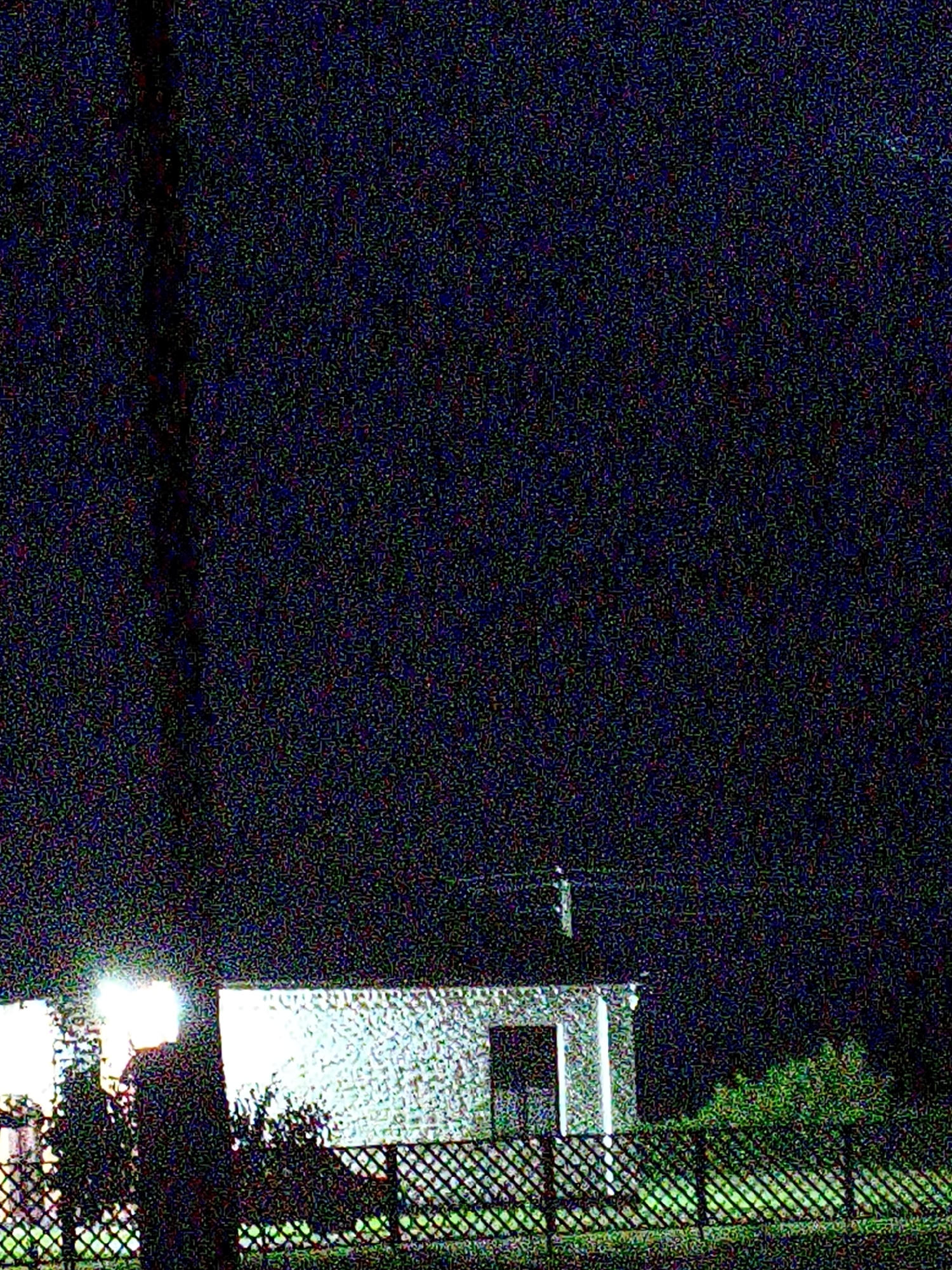 The image depicts a nighttime scene outside a small, white building with a prominent, illuminated entrance. A person is standing to the left of the entrance, silhouetted against a bright light source, obscuring their features. The building appears to be constructed of white brick or stone and has a tiled roof. In front of the building, a black metal fence with a diamond pattern encloses the area. To the right of the building, there is a dense cluster of green foliage. The overall environment suggests a residential or perhaps a small commercial property on the outskirts of a rural settlement, likely Ustyvytsia, Ukraine. The night sky is dark and speckled with what appears to be digital noise, common in low-light photography.