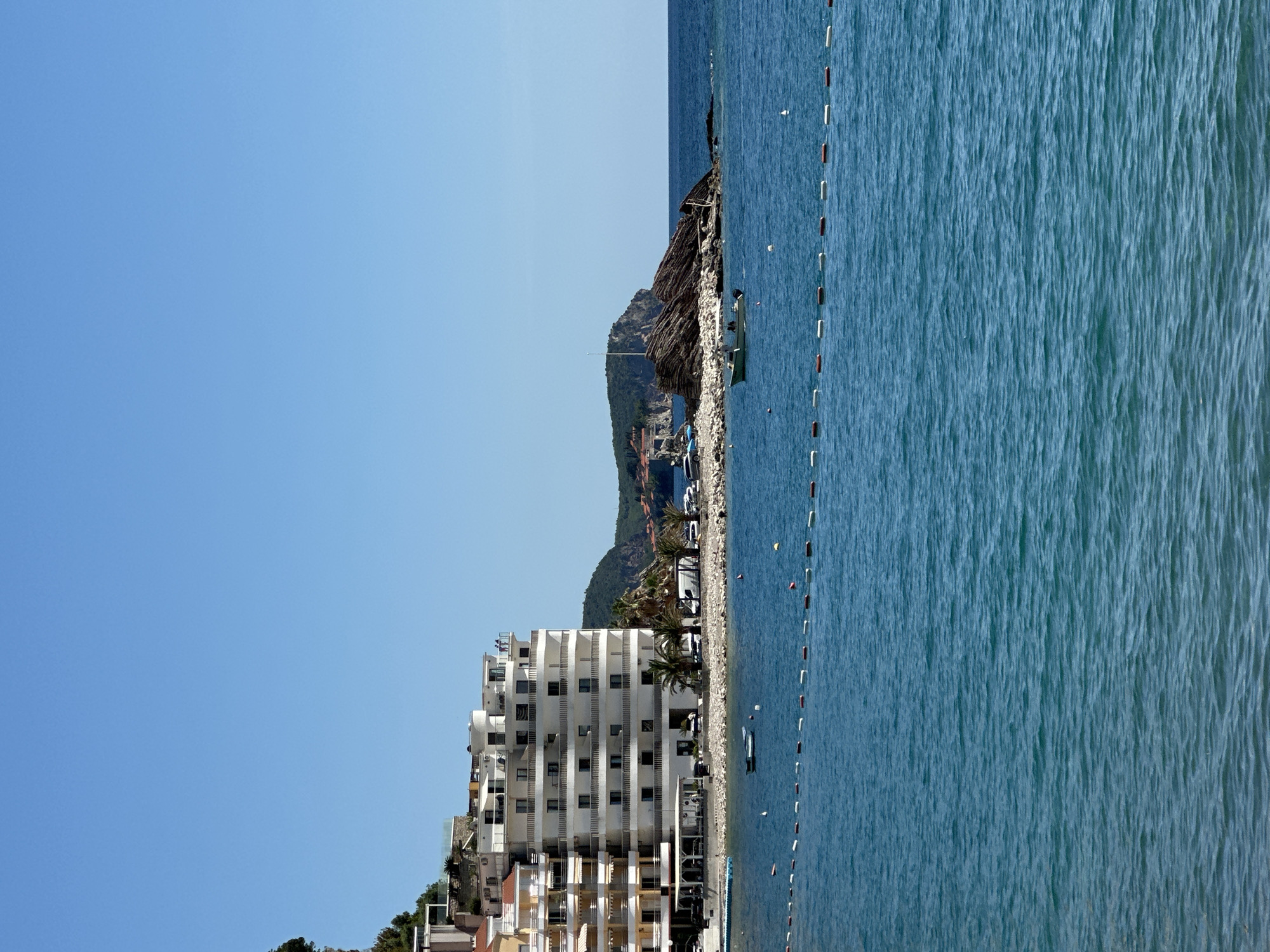 A seaside resort scene in Rafailovići, Montenegro, shows the Adriatic sea and beach on the right, extending vertically with small waves visible. The beach is marked off with a string of red and white buoys.
On the left, a multi-story white hotel with multiple balconies faces the sea. A row of smaller buildings is nestled below the hotel. Behind the hotel and buildings, a green, tree-covered mountain rises, leading to a rocky outcrop with some vegetation.
The sky is a clear, bright blue. Several parked cars can be seen between the hotel and the beach. A few palm trees are interspersed among the buildings and beach. A boat is floating on the water.