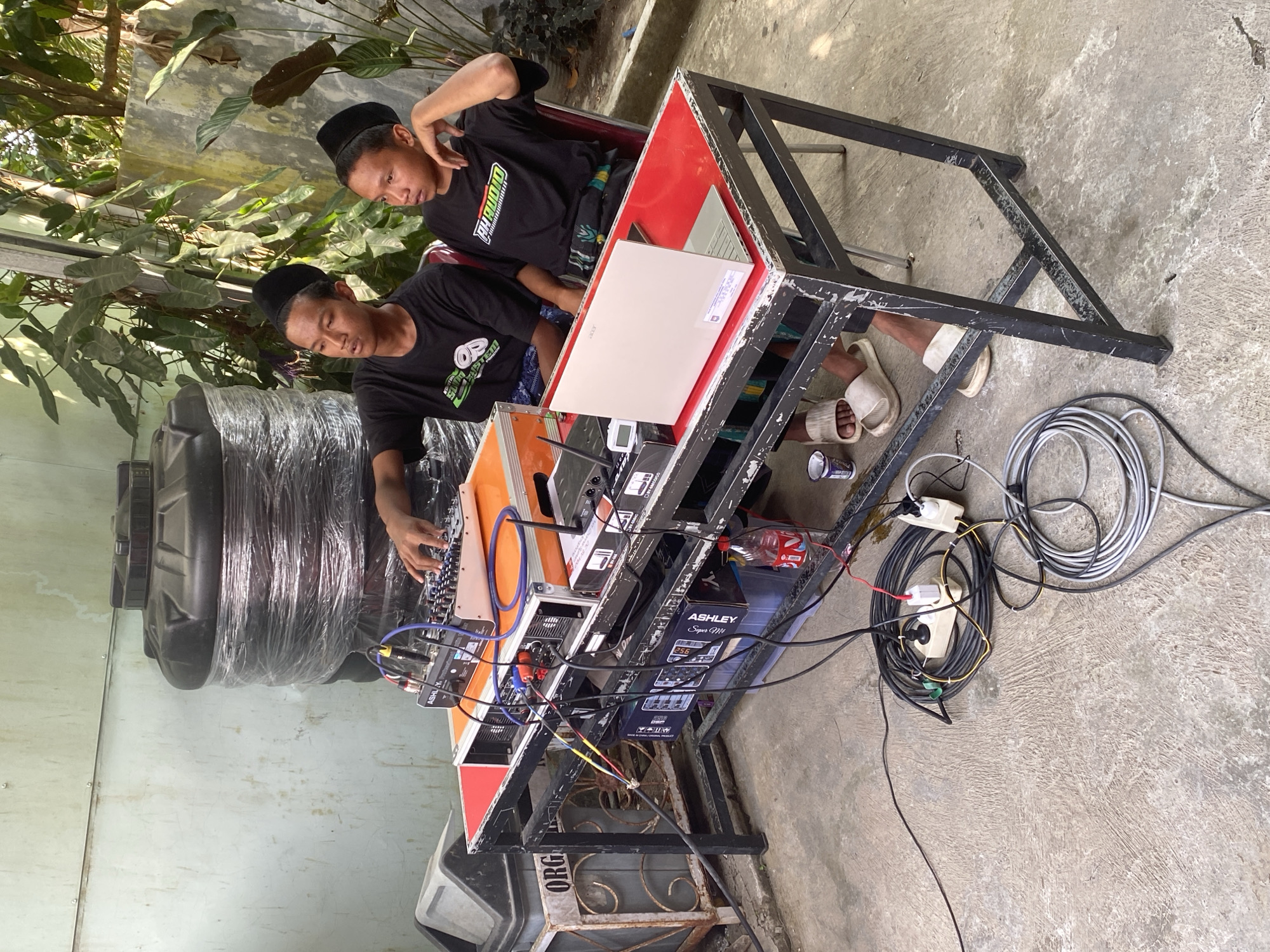 Two young males wearing black peci hats and black t-shirts are operating a DJ setup outdoors. The setup includes a mixing board, amplifiers, a laptop, and various cables and power strips, all placed on a red-topped metal table. The background features a large black water tank, a metal trash bin with the partial word "ORG" visible, and lush green foliage. The ground is paved with concrete. The scene appears to be in Gumelar Kidul, Indonesia, with the architectural style and vegetation being characteristic of the region. The overall impression is one of a makeshift outdoor event or gathering.