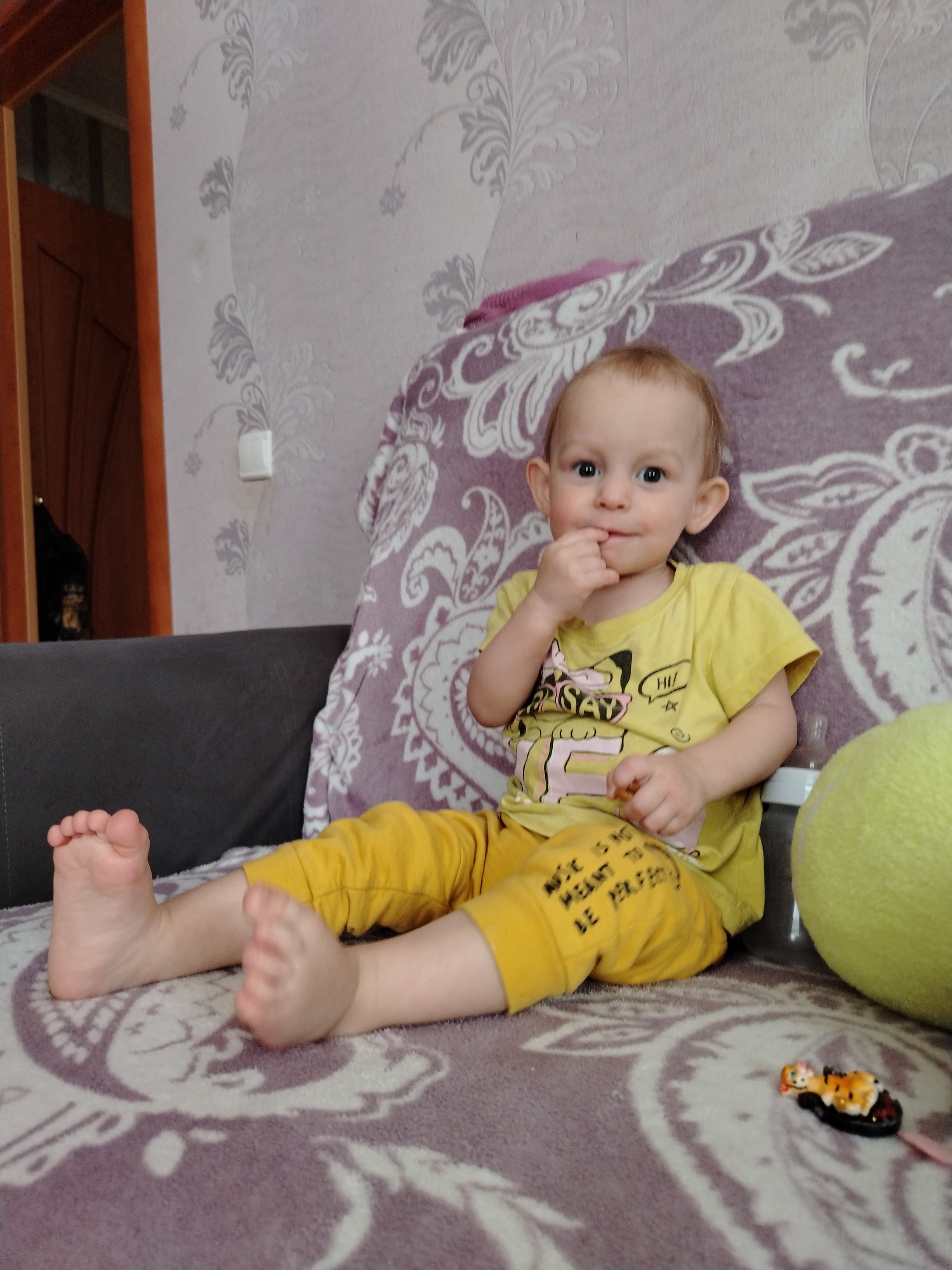 A young child is sitting on a couch, possibly in a home interior, with a patterned blanket. The child is wearing a yellow shirt and pants. The location is potentially in Nizhnyaya Gar', Russia. The child has a finger to their mouth and is looking at the camera. Next to the child is a bottle, a green ball, and a small toy. A grey sofa back is also visible. The wall behind the couch has a floral pattern. A partially-visible door is in the background on the left.