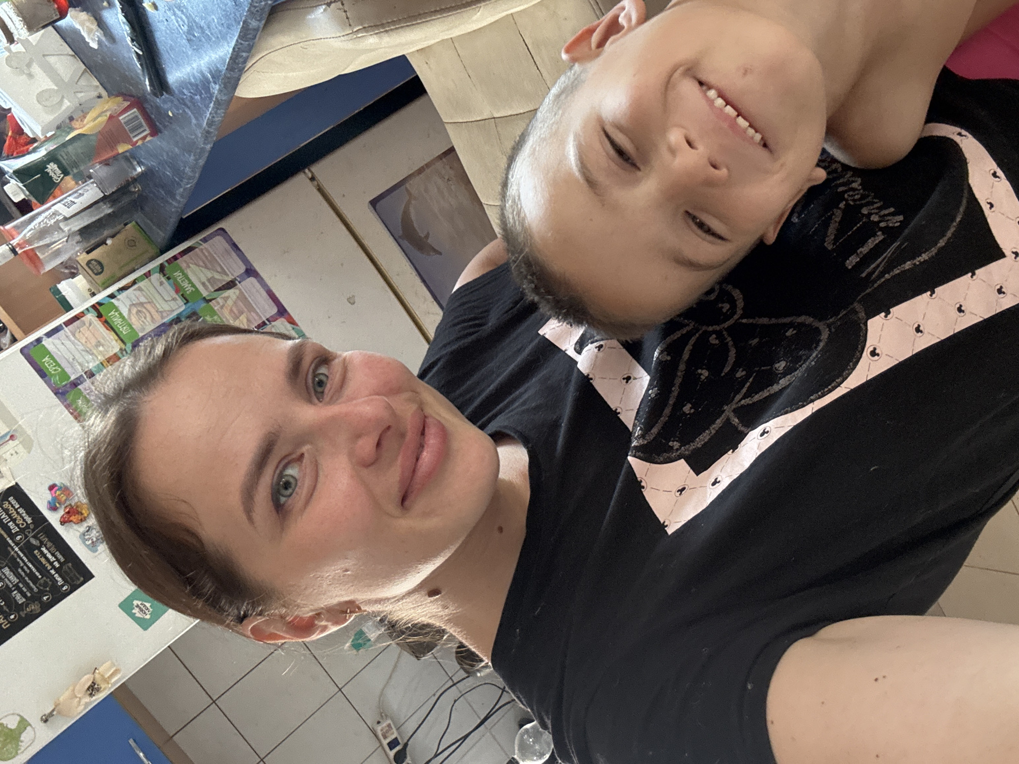 A close-up, selfie-style, over-the-shoulder shot features a woman and a young boy. The woman, with fair skin, light brown hair pulled back, and green eyes, is positioned in the foreground, looking directly at the camera with a smile. She is wearing a black t-shirt with a stylized pink and white design. The boy, with short brown hair and fair skin, is nestled close to the woman, also looking at the camera with a wide, toothy smile. He appears to be shirtless or wearing a dark garment. The background shows a kitchen environment with a refrigerator adorned with magnets and papers, and a countertop with various items, including juice cartons and bottles. A framed picture of a dolphin is visible in the upper right background. The overall impression is a candid, domestic moment. The location is specified as Rusalevka, Russia.