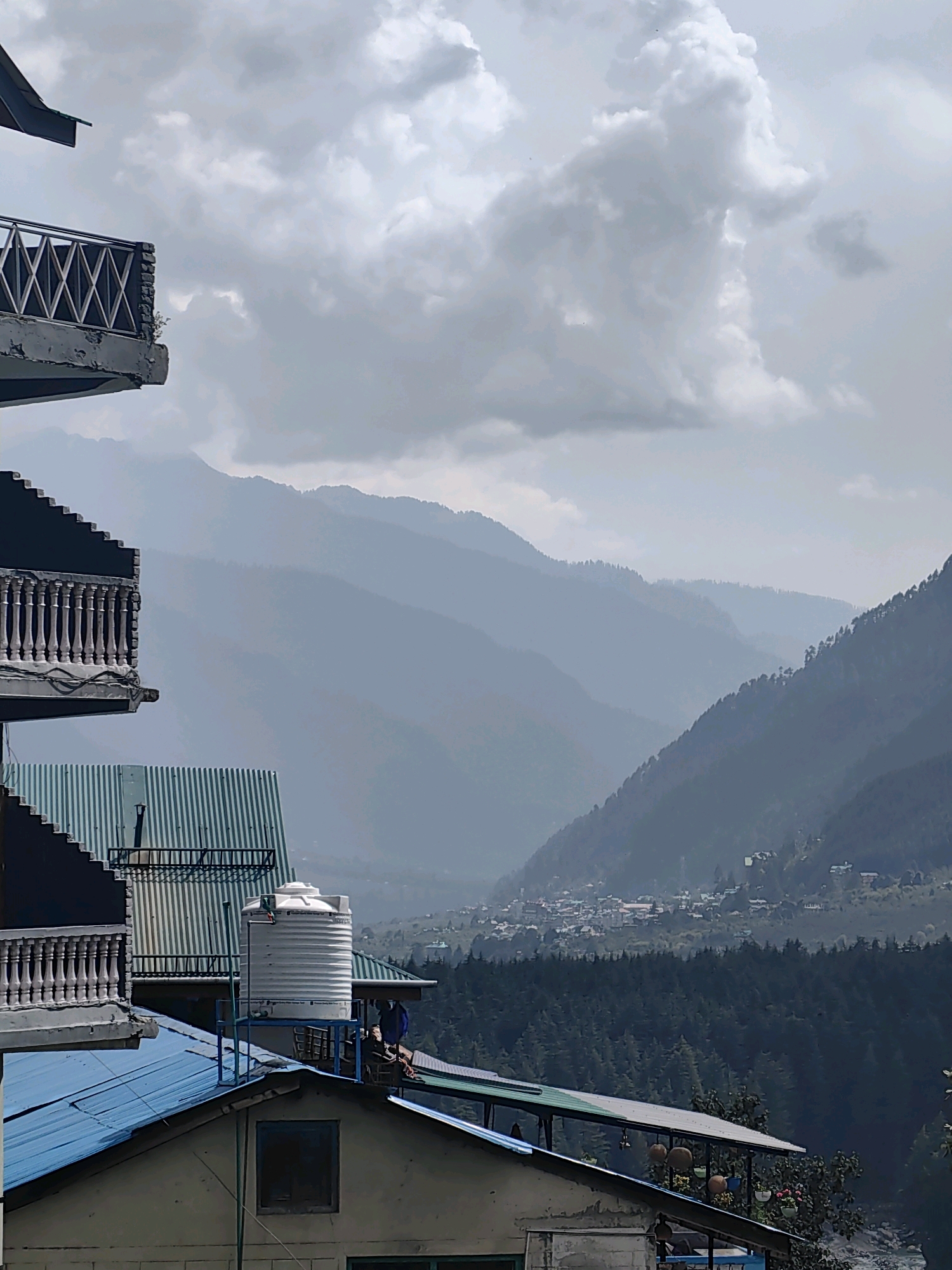 The image depicts a layered landscape with a focus on buildings in the foreground and a vast mountain valley in the background, characteristic of Vashisht, India. In the immediate foreground, a light blue metal roof of a building is visible. Above this, a section of a light-colored building with a dark-rimmed window is present. To the left and extending upwards, balconies with white ornate railings are stacked, attached to a grey structure. One of these balconies has a railing with a diamond pattern. A large white water tank sits on a metal platform on top of a green corrugated metal roof. Behind the buildings, the landscape transitions to a dense, dark green coniferous forest that carpets the lower slopes of the mountains. Further into the distance, hazy, layered mountains fade into the sky, with a small town or village nestled in the valley. The sky above is filled with dramatic, grey clouds, suggesting an overcast or potentially rainy day. Two individuals are partially visible in the middle ground, one standing near the water tank and another sitting. The overall scene conveys a sense of elevation and remoteness within a mountainous region.