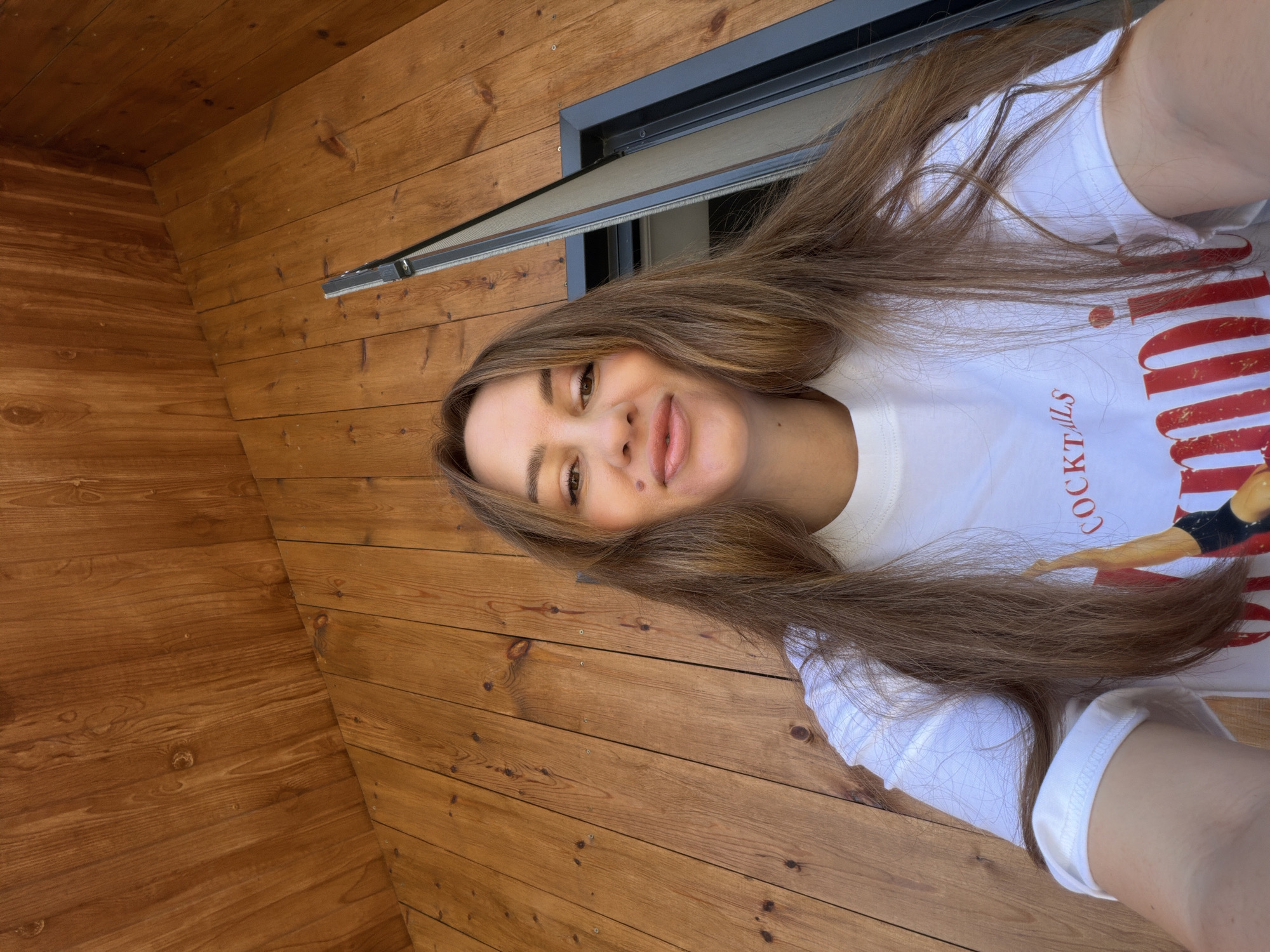 In the city of Malinovka, Russia, a woman with long brown hair, wearing a white t-shirt with red lettering, takes a low-angle selfie outdoors. The background consists of a wooden wall and a partially visible window frame. The woman's expression is a slight smile.