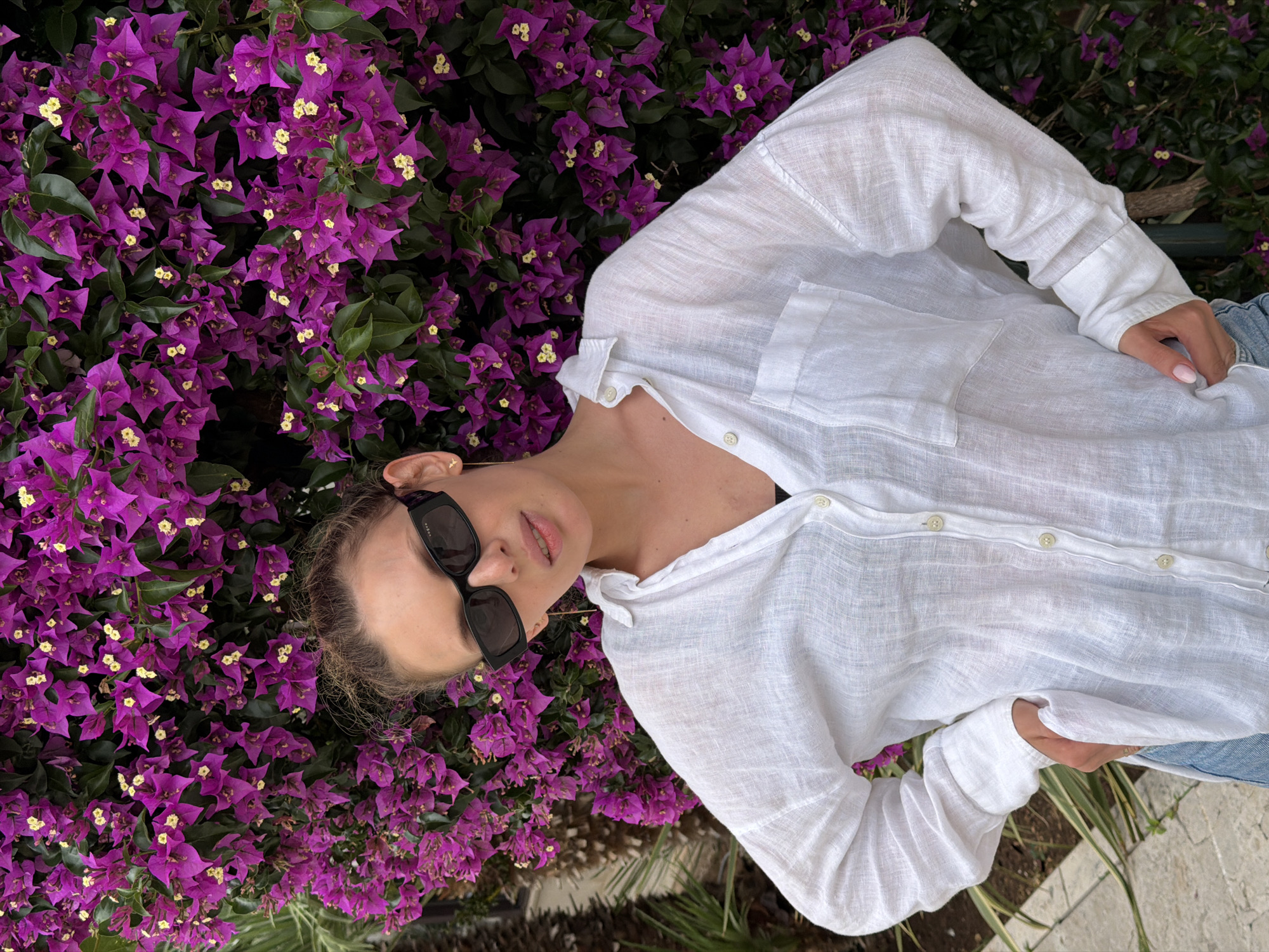 A woman is depicted in a close-up, rotated image, appearing to lie among vibrant purple bougainvillea flowers with green leaves, suggestive of a warm, Mediterranean climate like Budva, Montenegro. She is wearing a loose-fitting, white linen collared shirt, unbuttoned slightly at the top, and dark, rectangular-framed sunglasses. The word "GUCCI" is visible on the arm of her sunglasses. Her hair, appearing light brown or blonde, is pulled back.
Her left hand, with light pink manicured nails, is visible holding part of her shirt or resting near her hip, with a light blue fabric, possibly denim jeans, visible underneath. A small, subtle ring is on one of her fingers. Her posture appears relaxed, with her head slightly tilted and her lips softly parted.
The bougainvillea flowers dominate the background, showcasing their bright purple bracts and small, cream-colored true flowers. The lighting suggests it is daytime, with bright, diffused light highlighting the textures of her shirt and the flowers. The overall impression is one of a leisurely moment captured in a beautiful, natural setting.