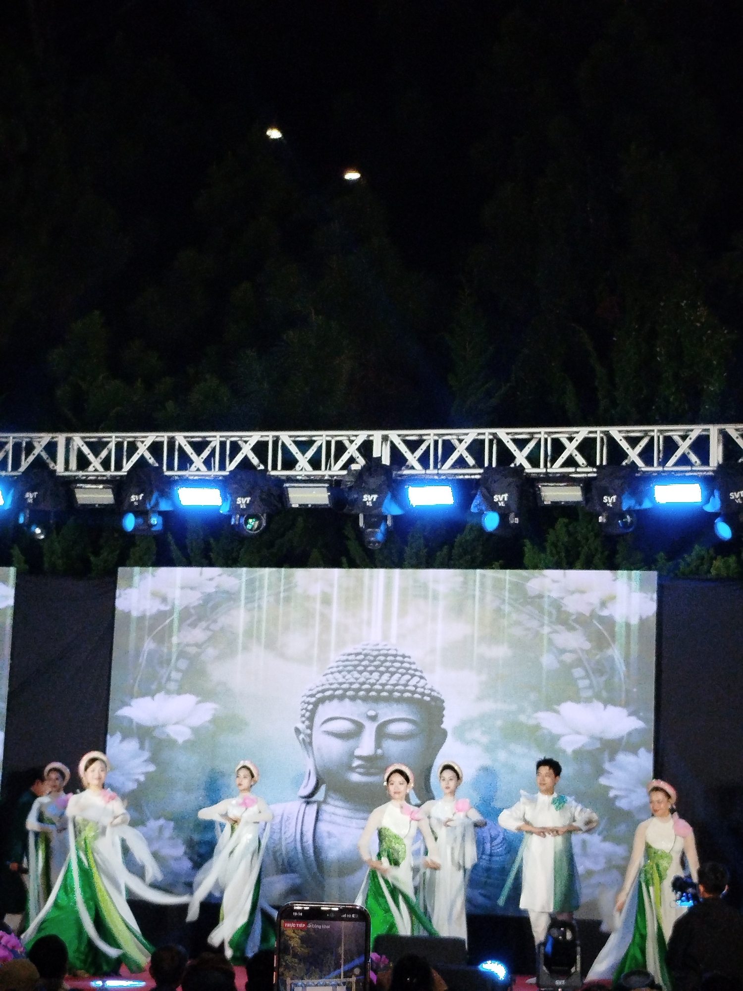 The image captures a performance in Da Lat, Vietnam. A group of dancers, attired in white and green traditional Vietnamese costumes, performs on a stage. Behind them is a large screen displaying a gray-scale image of a Buddha statue, surrounded by flowers. Above the screen, there's a truss structure fitted with several blue stage lights. The surrounding environment is dark, likely an outdoor setting, with trees visible in the background. Some audience members are seen in the foreground, along with a phone recording the performance. The lights above are illuminating the area.