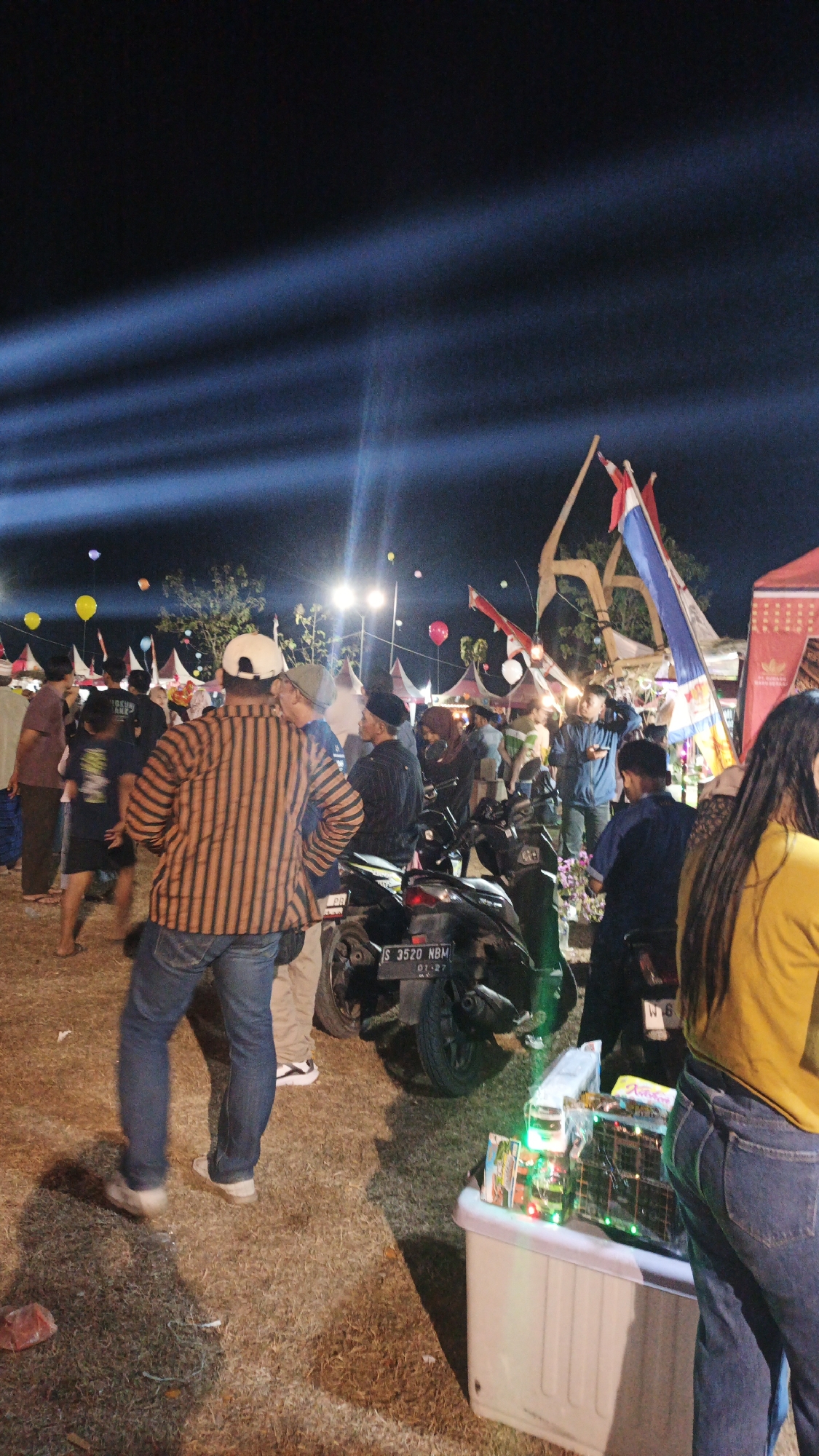 This image captures a nighttime scene at an outdoor event, possibly a festival or market, in Ketemasdungus, Indonesia. Several people are gathered, some appearing to be vendors and others attendees. In the foreground, a person wearing a striped shirt and jeans is facing away from the camera, looking towards a group of people. Behind them, several motorcycles are parked. Various stalls or tents, decorated with flags and lights, are visible in the background, suggesting a commercial or celebratory atmosphere. Several balloons are floating in the air, illuminated by stage lighting. The ground is covered in dry grass and some debris. The overall impression is of a lively, crowded event taking place after dark.