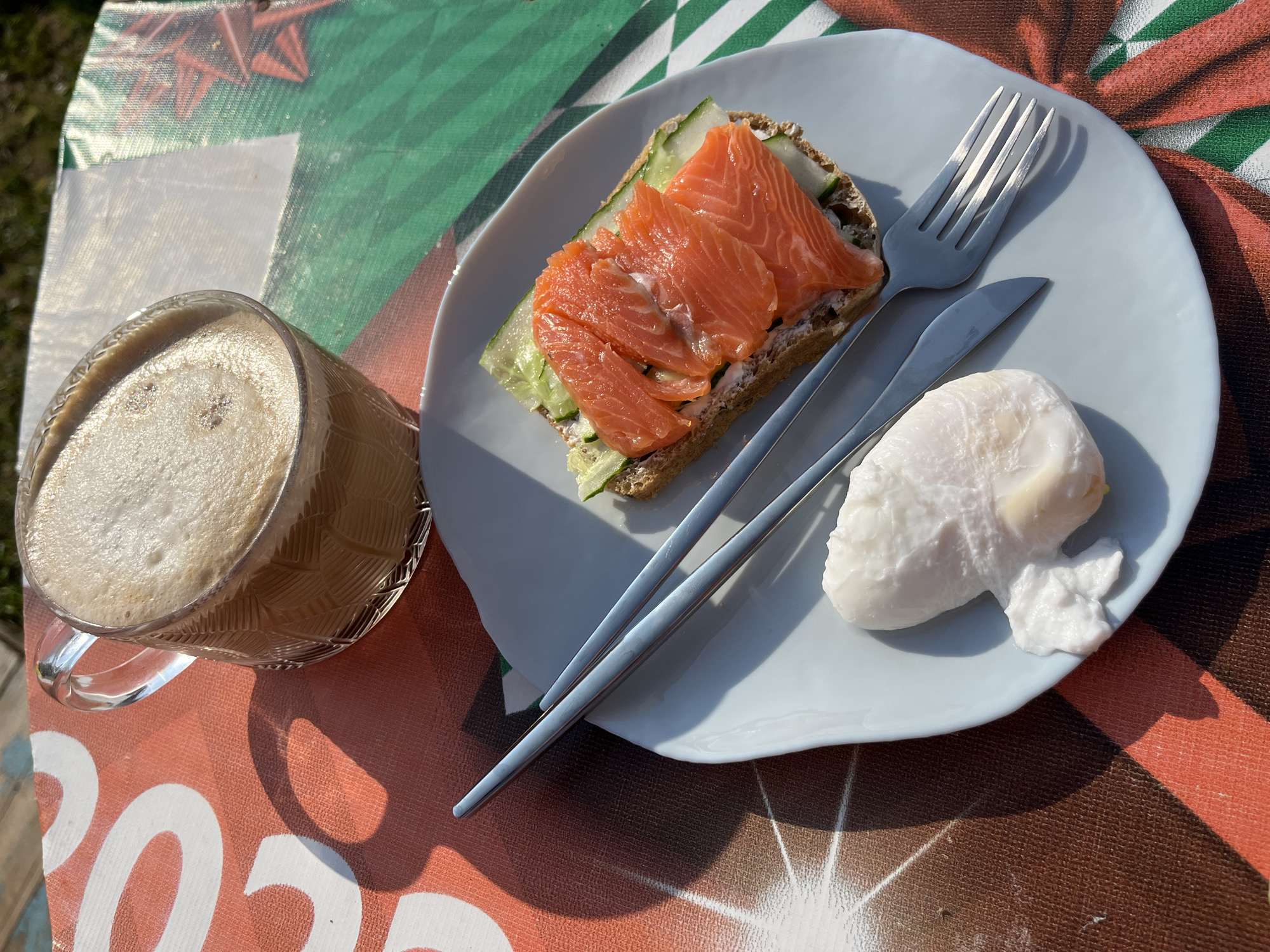 A breakfast spread is laid out on a patterned tablecloth in Chaikovsky, Russia. The subjects include a glass mug filled with a frothy beverage, a light blue plate containing a sandwich with smoked salmon and cucumber, and a poached egg. A fork and knife are also on the plate. The scene is set outdoors, indicated by the sunlight casting shadows and the presence of grass in the background. The tablecloth has a bold orange and white graphic design.
