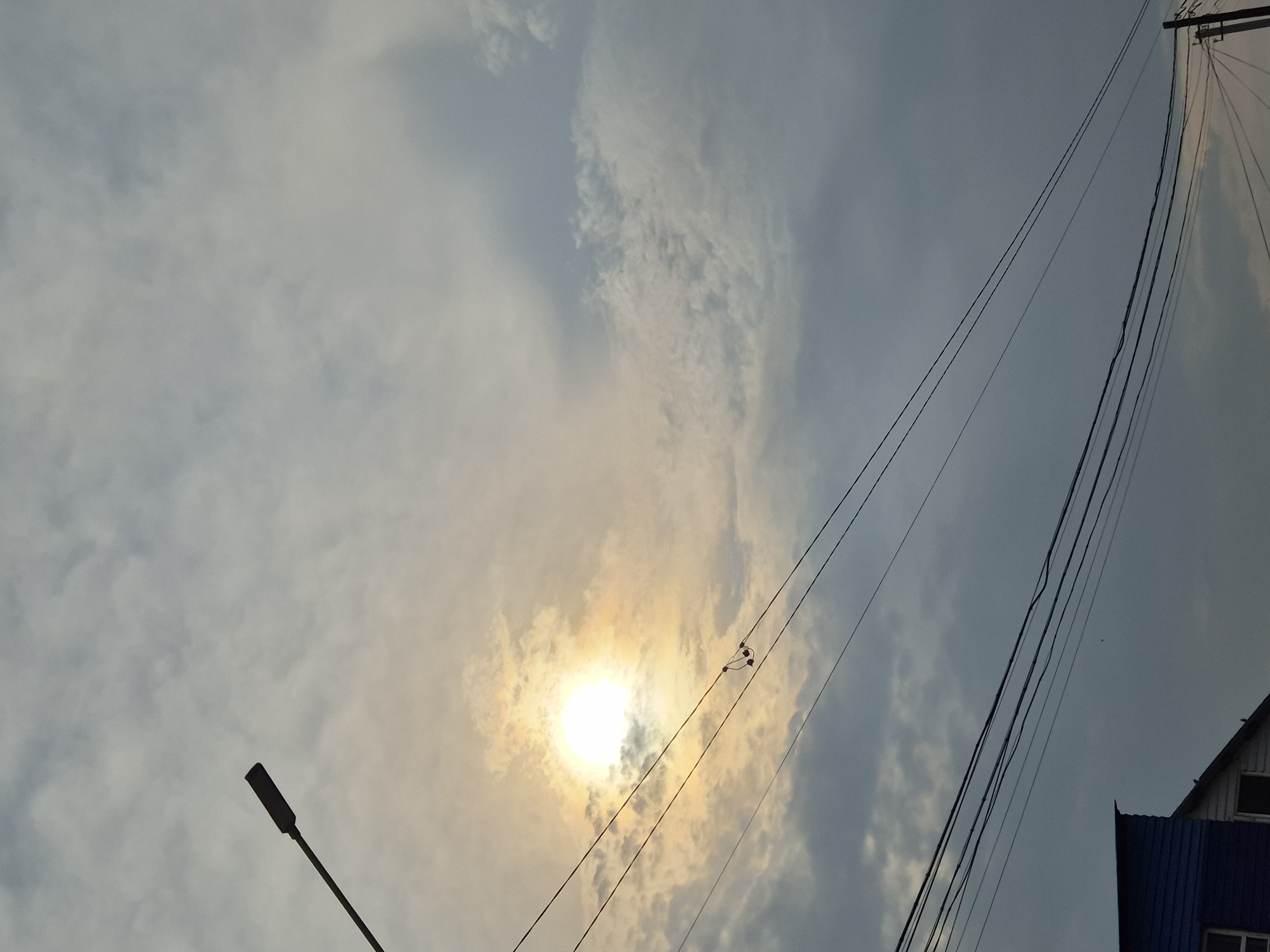 The image depicts a cloudy sky with the sun partially obscured by clouds, casting a bright glow. A street lamp extends from the left side of the frame, pointing upwards and to the right. Several power lines crisscross the sky, with a small object resembling earbuds hanging from one of the lines. In the lower left corner, the roof of a blue and white building is visible, suggesting an urban or suburban setting. The scene appears to be captured in Новоишимское, Kazakhstan. There are no people or explicit actions depicted.