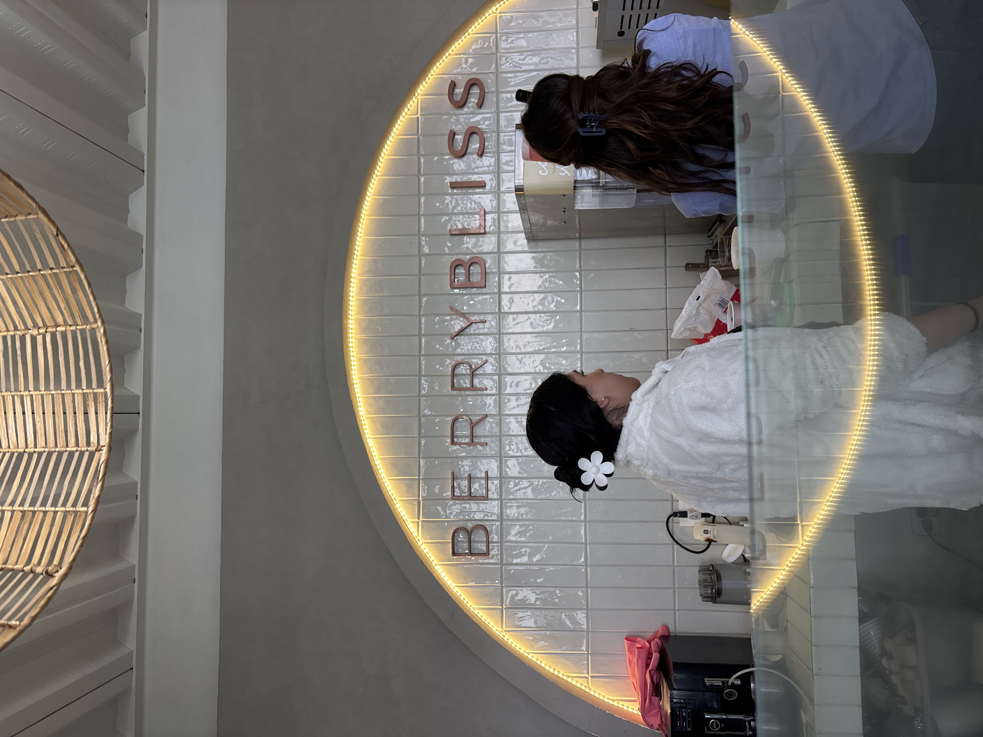 An indoor setting is presented with a tiled white wall featuring the vertically oriented word "BERRYBLISS" illuminated in red lettering and encircled by a yellow light. Two individuals are present; one is having their hair attended to by the other. The person receiving attention is wearing a white robe, while the other is dressed in a blue shirt. Miscellaneous items such as a dispenser and pink clothing are visible on the lower part of the wall, and a woven rattan basket is located on the side. The scene is in San Bernardino Tlaxcalancingo, Mexico.