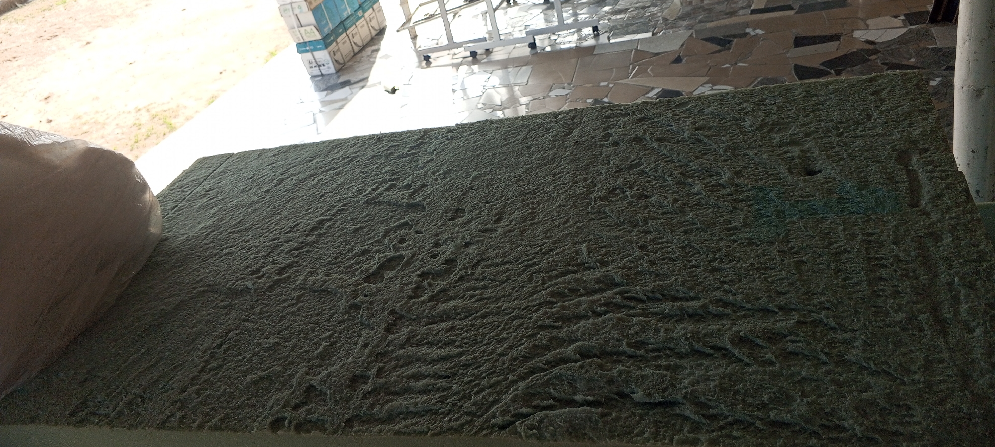 The image shows a close-up of a rough, textured, pale green material, possibly insulation or foam, covered in dust or debris. The material is laid out horizontally in the foreground, filling most of the frame. To the left, a large, soft, cream-colored object, possibly wrapped in plastic, partially obscures the edge of the green material. In the background, a tiled floor with irregular, multi-colored geometric tiles is visible, along with a white pillar to the right. Sunlight streams in from the left, casting shadows and highlighting the texture of the material. Further in the background, stacks of white boxes, possibly paper, are visible, along with metal shelving and a large glass or plastic structure. The scene appears to be outdoors or in a semi-open space, possibly a construction site or storage area in Jalingo, Nigeria.