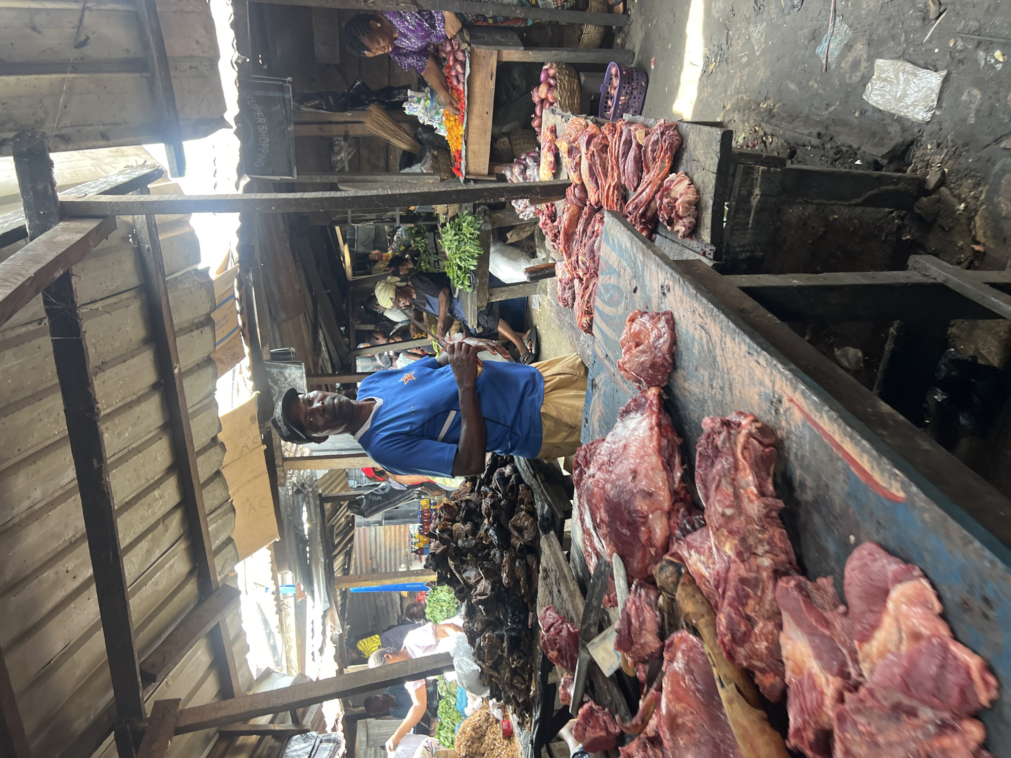 This is a photograph taken at a busy open-air market in Nkpor, Nigeria, likely a butchery section. In the foreground, a wooden table is laden with large cuts of raw red meat, some appearing to be beef or lamb, with visible fat and muscle. A knife rests on the table. A man wearing a blue t-shirt with a star emblem and a black baseball cap stands behind the table, holding a piece of meat. He is looking towards the camera.
In the background, more market stalls are visible, with awnings made of corrugated metal and wooden structures. Other individuals are present, engaged in market activities. To the right, a woman in a purple garment is seen arranging colorful vegetables, possibly peppers and onions, on a stall. Baskets containing produce and onions are also visible. The ground is dirt and appears somewhat cluttered with debris. The overall impression is of a vibrant, active marketplace environment.