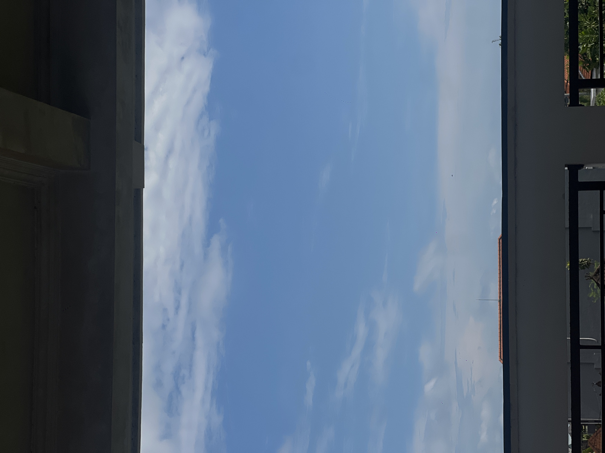 The image shows a view of the sky with scattered clouds and a blue hue, framed by architectural structures. The left side of the frame features a portion of a building with a beige color. On the right, there is a building with a white wall and a black iron fence, with hints of greenery visible through the fence. In the sky, a series of tiny dots may represent birds flying in formation. The location could be Canggu, Indonesia, but there are no specific local details to confirm. The scene is static and depicts an outdoor environment.