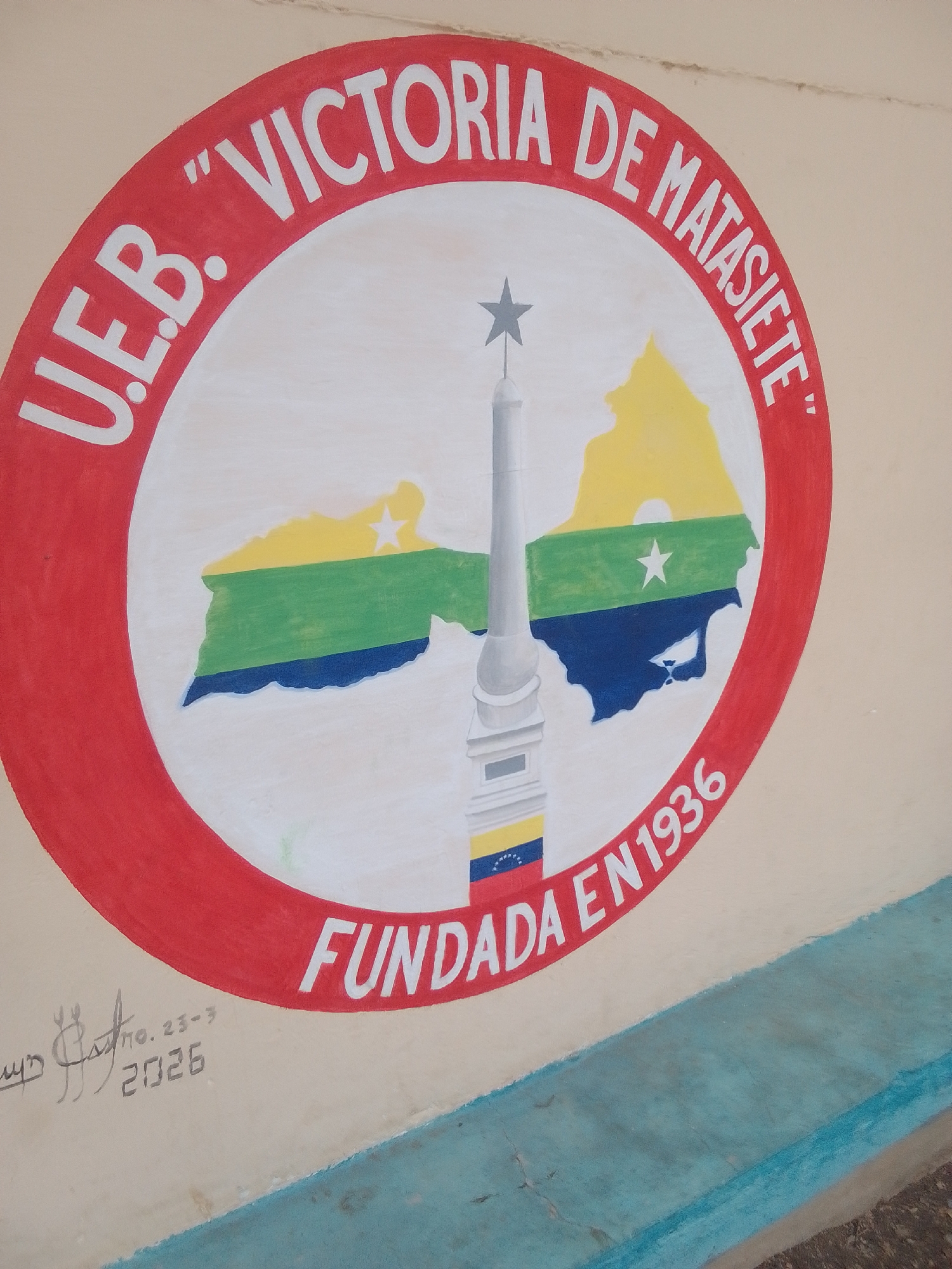 This is a photograph of a mural painted on a wall, likely outdoors. The mural is circular and features a coat of arms or emblem. At the top of the circle, in white capital letters against a red background, is the text "U.E.B. 'VICTORIA DE MATAS." Below this text, within the circle, is a stylized depiction of the Venezuelan flag colors (yellow, green, and blue) overlaid on a map-like shape. In the center of this map shape is a white star. A tall, grey, obelisk-like monument with a star on top stands in front of the map, and a section of the Venezuelan flag is painted on its base. Along the bottom of the circle, in white capital letters, is the text "FUNDADA EN 1936". To the left of the mural, there is a handwritten signature or inscription with the date "2026". The wall appears to be painted a light beige color, and a blue stripe runs along the bottom of the frame. The lighting suggests it is daytime, and the setting seems to be a public space in La Tagua, Venezuela, given the context.