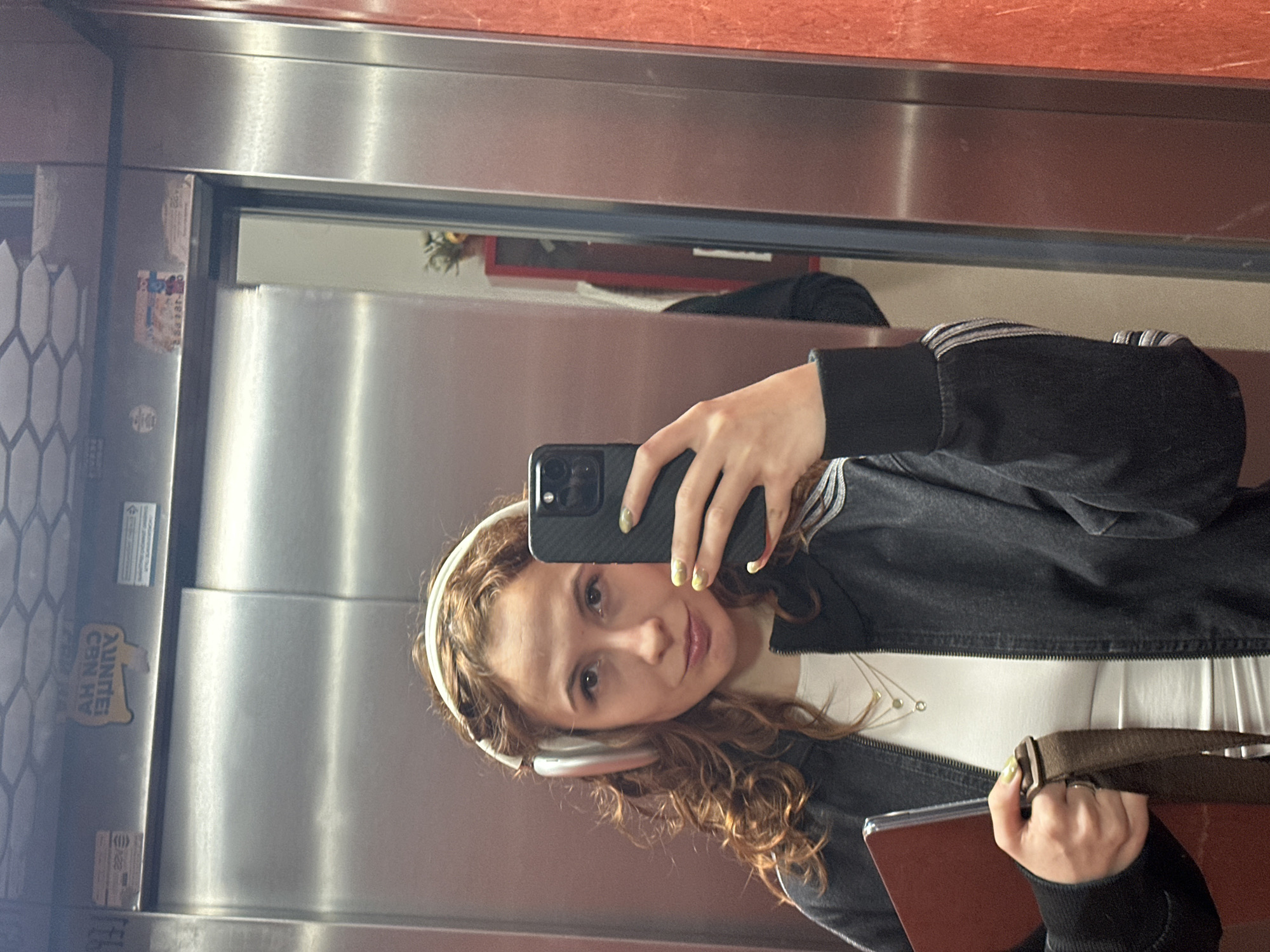 A young woman is depicted taking a mirror selfie inside an elevator in Novi Sad, Serbia. She has long, curly brown hair and a fair complexion, looking directly at the camera with a subtle, pleasant smile. She is wearing white over-ear headphones, a white top, and a dark jacket, possibly denim or canvas, which features light-colored stripes on the sleeves. Her nails are painted light yellow with a faint pattern.
She holds a dark smartphone in her right hand to capture the image. Her left hand is holding a brown strap, likely from a bag, and a reddish-brown object, possibly a book or a tablet. The elevator's interior consists of metallic, silver-toned walls, which appear to be stainless steel, and a reddish-brown panel along the top. The selfie is taken in a large mirrored section of the elevator wall. To the left, a wall panel with a hexagonal or honeycomb texture is visible.
Reflections in the mirror provide glimpses of a green object and a red frame in the background. On the left metallic wall, an inverted sticker is visible, partially reading "AUNTE! C8N H¥", with a small dog-like graphic beneath it. Several other smaller, mostly illegible labels and stickers are also present on the metallic surfaces. The indoor setting offers no clues regarding the time of day or weather.