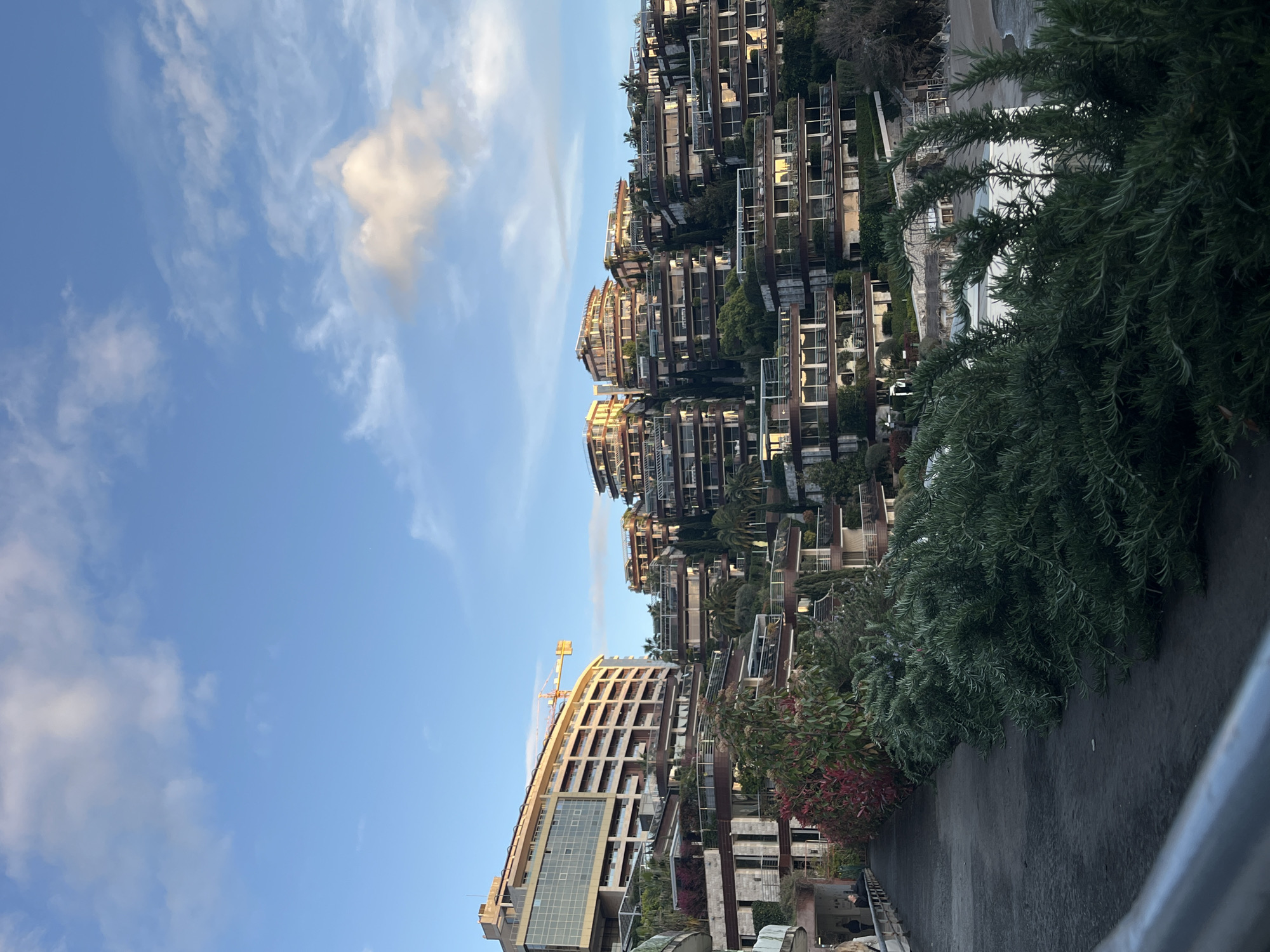 This image depicts a panoramic view of the Dukley Gardens complex and surrounding area in Budva, Montenegro, overlooking Bečići beach and the Adriatic Sea. The time of day is characterized by clear blue skies with scattered white clouds and low-angle golden sunlight, suggesting either early morning or late afternoon.
In the foreground, a dark, paved walkway curves right, with a metal railing partially visible on the far left. Two indistinct human figures, one in a dark jacket, are visible walking away from the camera on this path. The path is bordered by a line of red and green foliage. To the immediate right, dense dark green shrubbery, possibly rosemary, forms a prominent bush line, partially obscuring the lower right portion of the scene. Beyond this vegetation, a sandy beach and the calm waters of the Adriatic Sea are visible, along with white, low-rise beach structures.
The midground and background are dominated by a series of terraced, modern architectural structures built into a steep, vegetated hillside. On the far left, a large, multi-story building with a curved, light-colored facade and numerous balconies reflects the golden sunlight. A yellow construction crane is visible behind its upper section, indicating ongoing work. To the right of this building, numerous multi-level apartment or villa-style buildings, characteristic of Dukley Gardens, cascade down the hillside. These structures feature extensive balconies with glass railings, modern facades, and integrated Mediterranean vegetation, including palm trees and various shrubs. The buildings are bathed in warm, golden light, creating a strong contrast with areas in shadow.