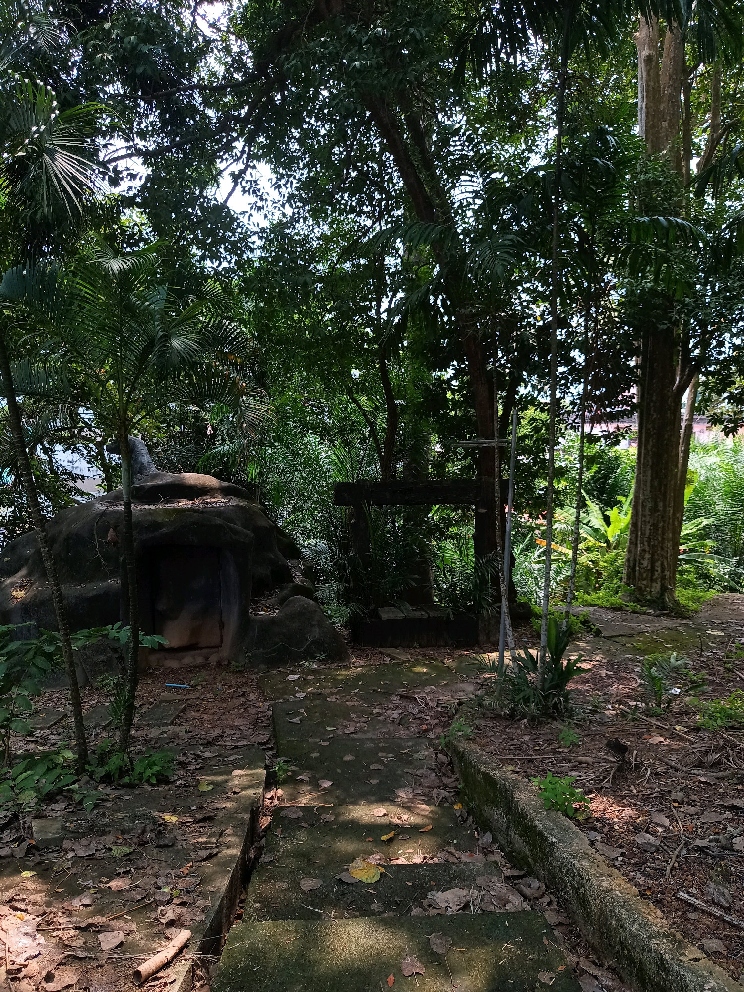 The image depicts a pathway in an outdoor environment, possibly within a park or jungle area, located in Sai Thai, Thailand. The pathway, made of concrete slabs, is partially covered with fallen leaves and is bordered by overgrown vegetation. To the left of the pathway, there is a large rock formation that appears to be artificial, with a dark opening resembling a cave entrance at its base. A tall palm tree stands to the left of this rock. Further into the scene, to the right of the pathway, a wooden structure resembling a gallows or frame is visible, with vertical poles and a horizontal beam. The background is dense with lush green foliage, including large trees with thick trunks and various types of plants, suggesting a tropical environment. The lighting indicates it is daytime, with sunlight filtering through the canopy and casting shadows on the pathway. No people are visible in the image.