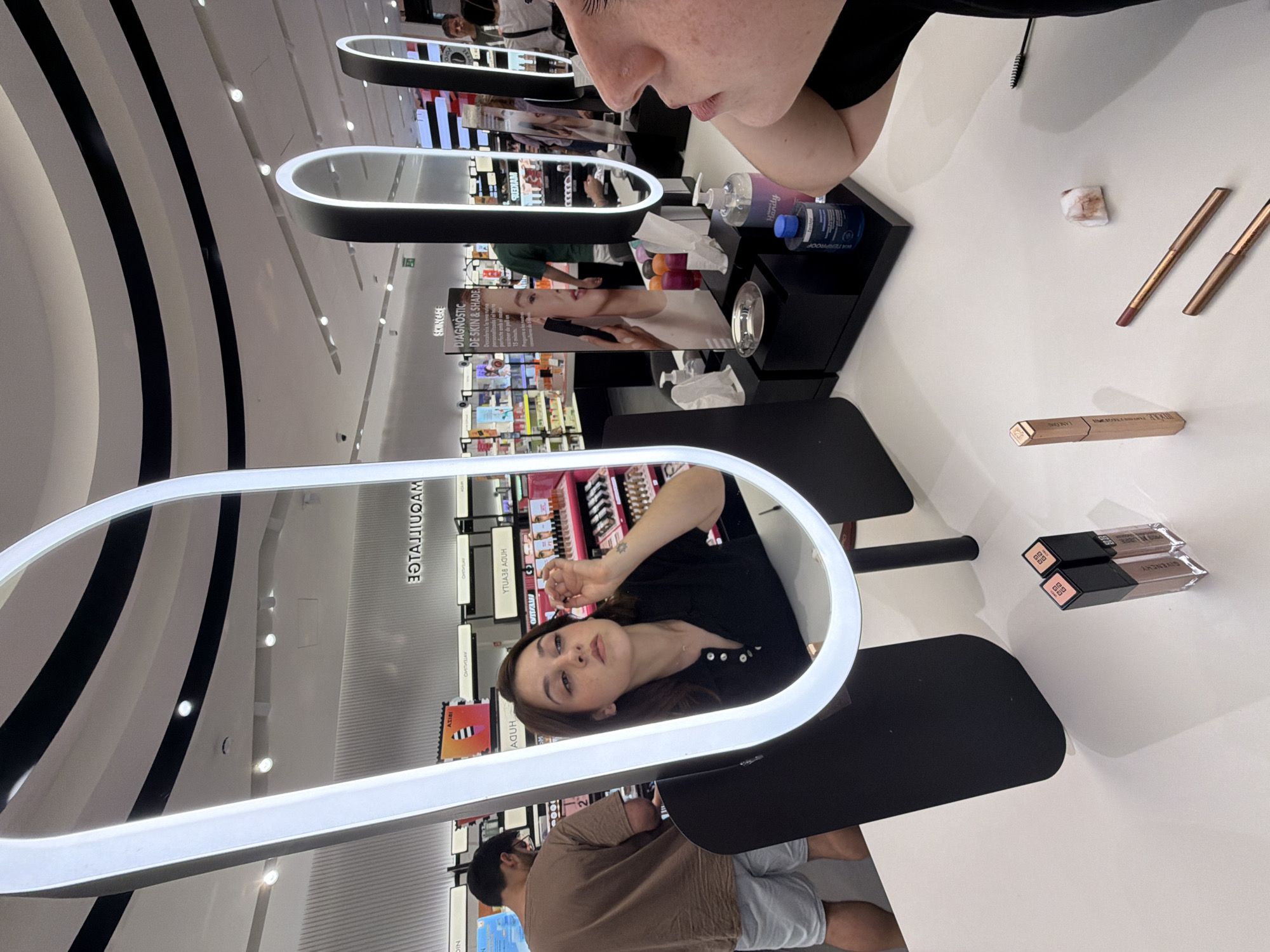 A woman is applying makeup in front of a large illuminated mirror in a retail store. She is positioned in the center of the frame, reflected in the mirror, and is focused on applying product to her eye. She has brown hair and is wearing a dark collared shirt. To the left of the woman, a man wearing a tan t-shirt and light shorts is visible from the back, appearing to be shopping. In the foreground, on a white counter, are several makeup products including two tubes of lipstick, a tube of mascara, and two makeup pencils. Behind the woman, shelves filled with various beauty products are visible. The store environment appears clean and well-lit, with a modern design. The woman has a small tattoo on her wrist. The reflection of the store's signage is visible in the mirror, with some text appearing backward. The overall scene suggests a makeup counter within a department store or beauty retailer. The image was taken in Barcelona, Spain, at a Sephora store in the El Corte Inglés department store located on Passeig de Gràcia.