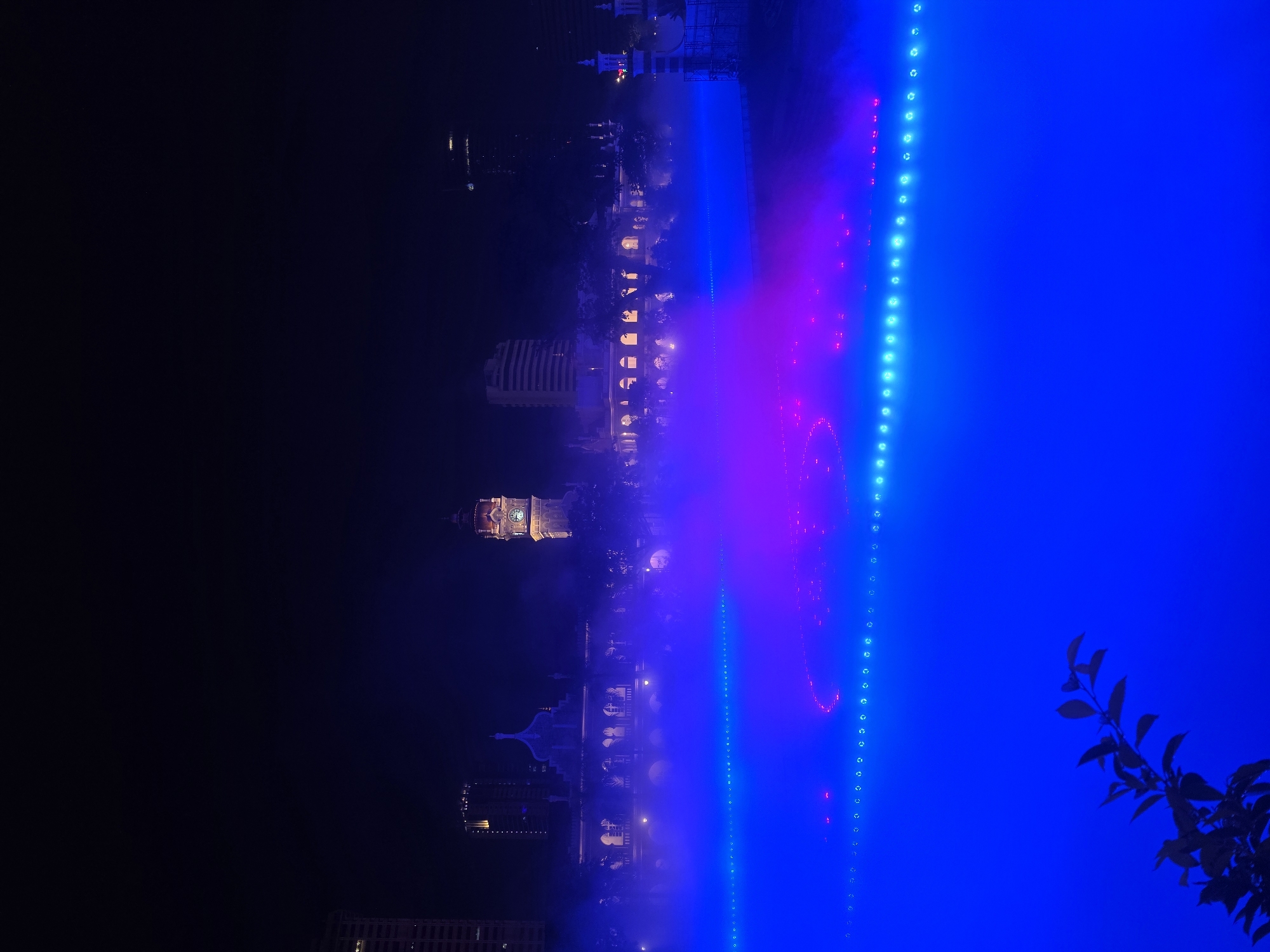 The image depicts a night scene in Kuala Lumpur, Malaysia, featuring the Sultan Abdul Samad Building and surrounding illuminated water features. The Sultan Abdul Samad Building, characterized by its Moorish architecture, is visible in the background, its clock tower illuminated. In the foreground, a water feature displays a dynamic light show with shades of blue and pink. A vertical line of lights is located on the right side of the image. Silhouetted foliage occupies the lower right corner. Other buildings are faintly visible in the background.