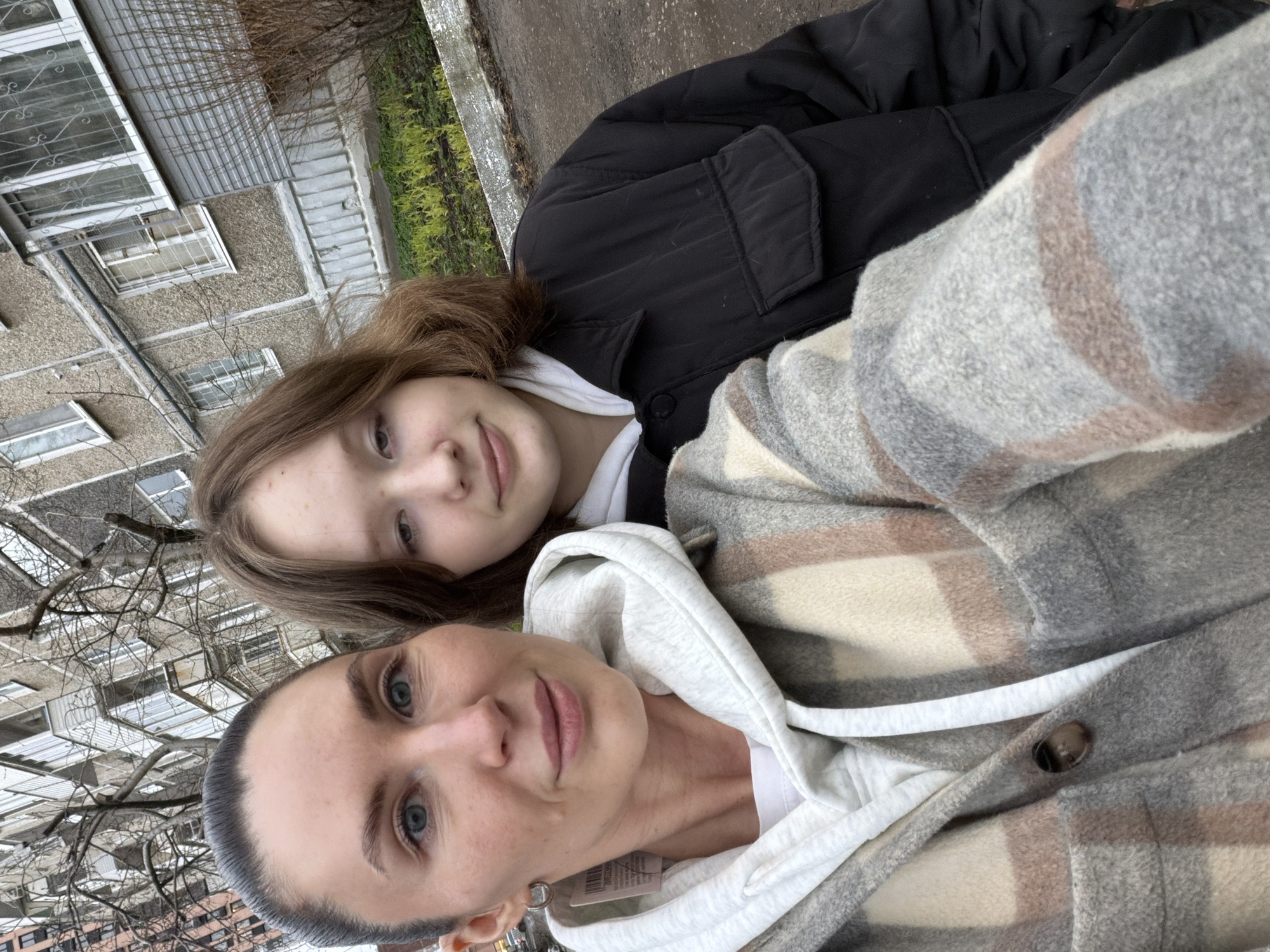 The image is a selfie taken by an adult woman with a younger female, possibly a teenager. The woman is in the foreground, wearing a grey and beige plaid jacket over a white hoodie. She has short, dark hair and fair skin. The younger female is behind her, wearing a black jacket over a white top, with shoulder-length brown hair. They are both looking at the camera.
The background suggests an urban environment, likely in Perm, Russia. An apartment building is visible, with multiple windows. Bare tree branches are also noticeable, suggesting it may be late autumn or winter.
