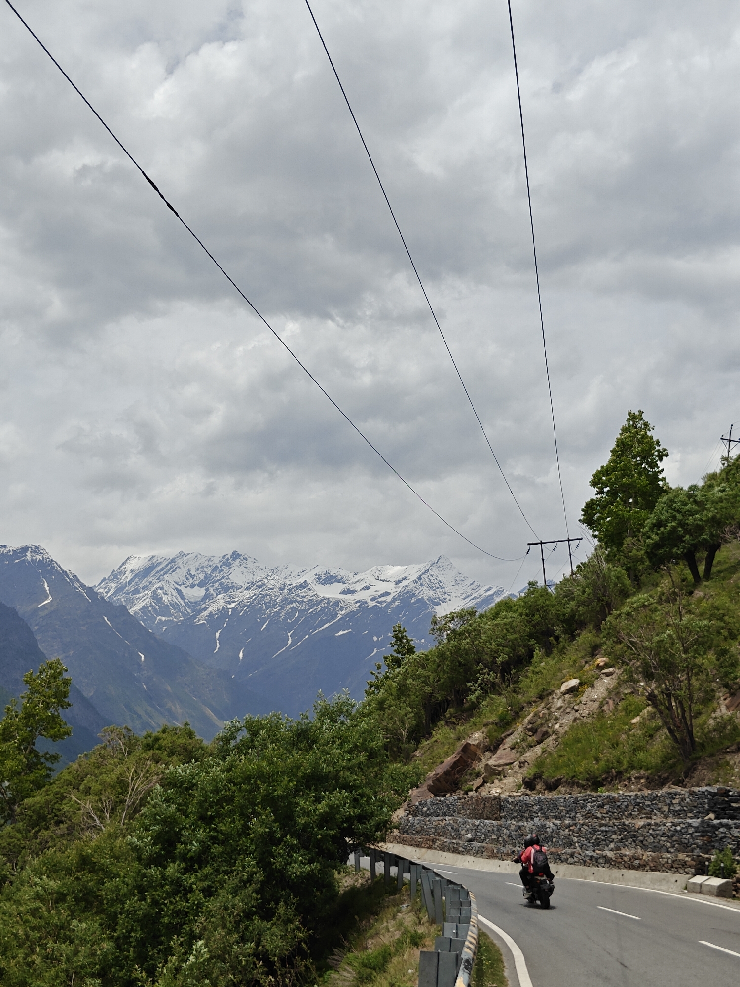 A motorcycle rider wearing a red jacket and a backpack is seen from behind, driving on a winding asphalt road. The road is bordered by a metal guardrail and a stone wall on the right. The road cuts through a lush green mountainous landscape. In the background, snow-capped mountains rise against a cloudy sky. Overhead, power lines stretch across the frame, supported by utility poles. The scene appears to be located on a mountain pass, possibly in the region of Jagle, India, given the presence of snow-capped peaks and dense vegetation.