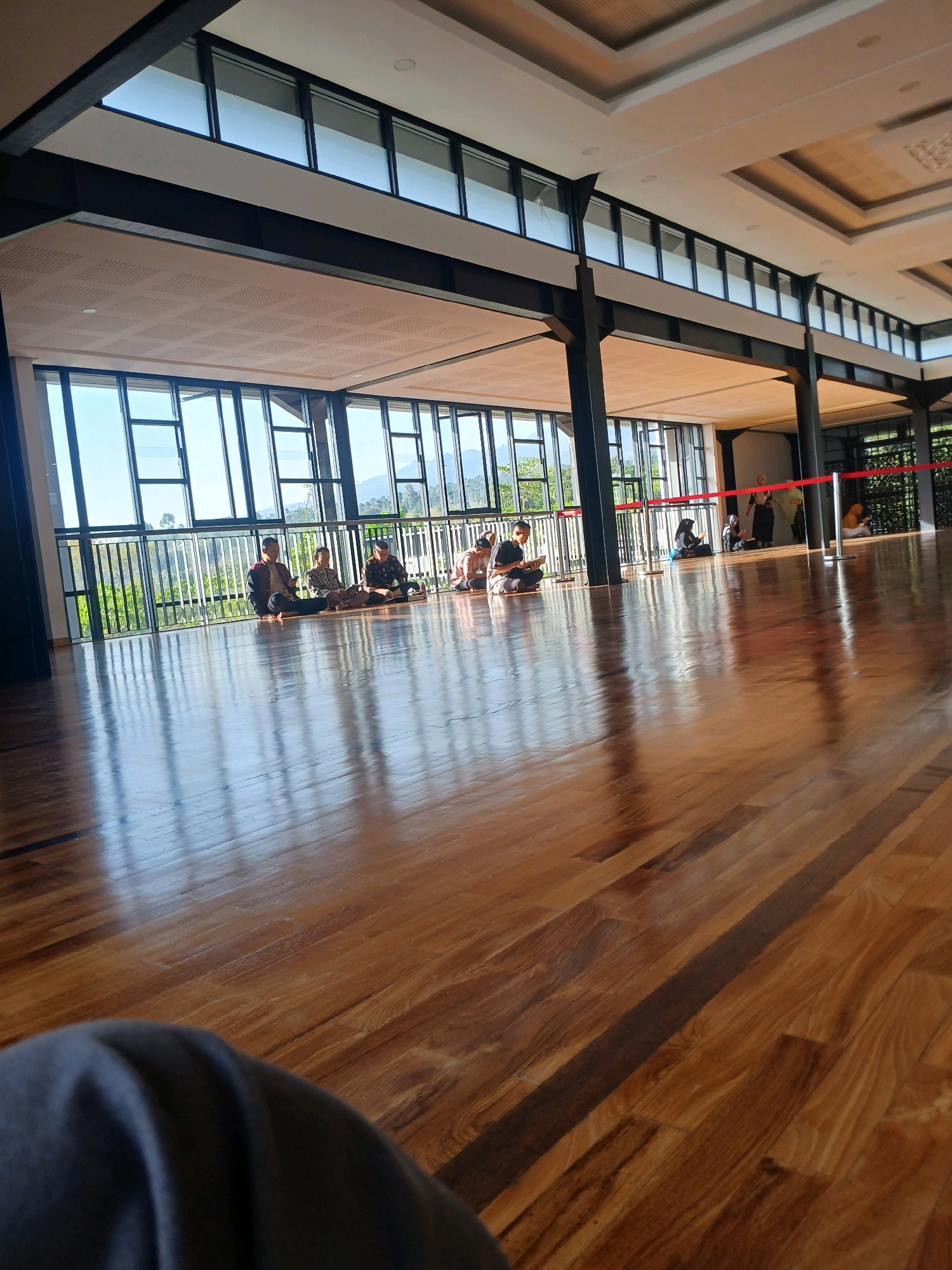 The image depicts a spacious, indoor environment with a polished wooden floor reflecting the natural light streaming through large windows. The windows offer a view of lush greenery and distant mountains, suggesting a scenic location. Several individuals are seated on the floor in various sections of the room, some appearing to be engaged in reading or quiet contemplation.
Specifically, on the left side of the room, a group of people are seated cross-legged. Moving towards the center, a man in a jacket and another individual are also seated on the floor. Further to the right, more individuals are gathered. In the background, near a red rope barrier, a few more people are visible, one appearing to be standing. The architecture features dark structural beams and a high ceiling with illuminated panels. The overall atmosphere suggests a tranquil and possibly educational or spiritual gathering space. The scene is located in Arjasari, Indonesia.