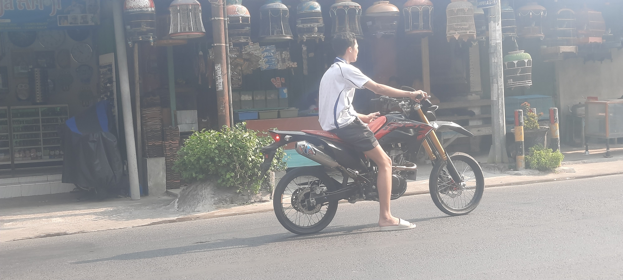 A young man is seated on a red and black dirt bike, facing left. He is wearing a white t-shirt with blue trim and black shorts. The motorcycle is parked on the side of a street in Selopuro, Indonesia, in front of a storefront. The storefront displays various birdcages hanging from the ceiling and various items on shelves inside. A large green bush is planted to the left of the motorcycle, and a black tarp covers another object to the far left. The street is paved, and the environment appears to be a market or commercial area.