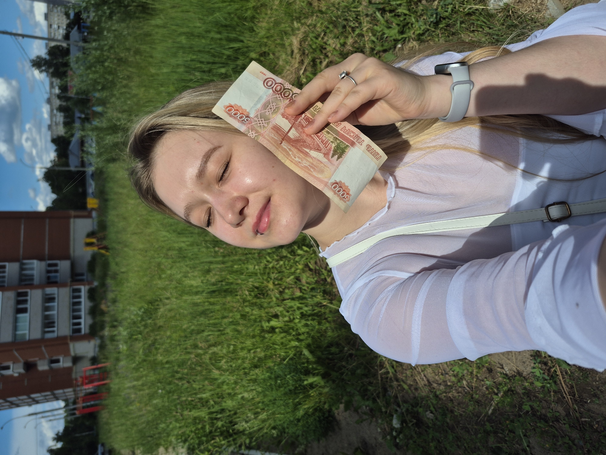 A young Caucasian woman with blonde hair and closed eyes holds a 5000 ruble banknote in her right hand. She wears a sheer white top and a silver ring on her left ring finger. A white strap from a bag is visible over her shoulder. Her left wrist has a gray fitness tracker. She stands outdoors in a grassy area with a brick apartment building and playground equipment in the background. The sky is blue with scattered clouds. The location is the city of Chaikovsky, Russia.