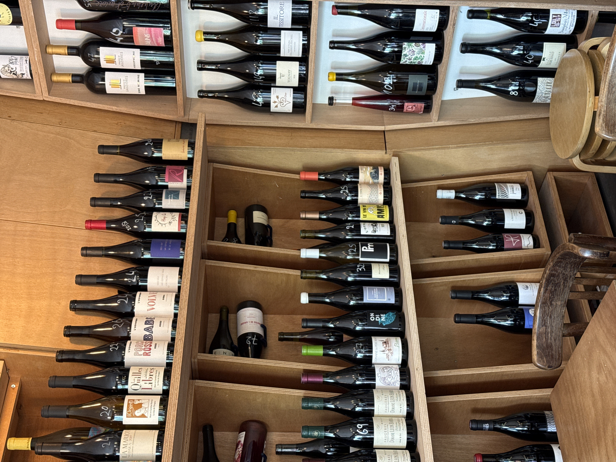 The image shows a wine rack filled with various bottles of wine. The bottles are arranged in wooden compartments, some standing up and others lying horizontally. The labels indicate that they are primarily French wines. The camera angle suggests the photograph was taken indoors. Some stacked wooden chairs are visible to the right of the wine rack. The context and label information suggest that the scene is in a wine store or cellar in Paris, France.
