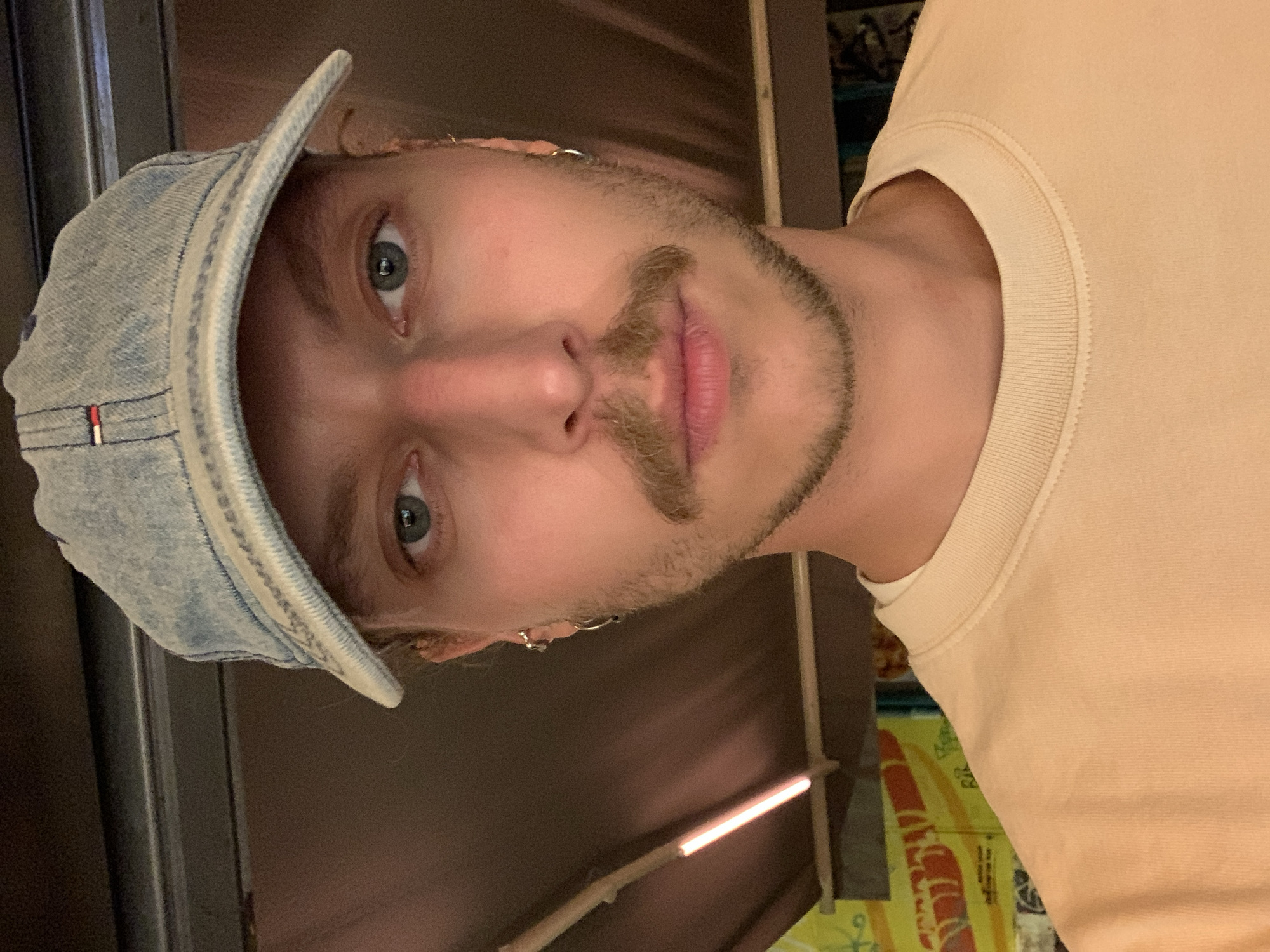 A person with light skin, light hair, a mustache, and a short beard is wearing a light-blue denim cap and a beige T-shirt. They appear to be indoors in a dimly lit place. There is no visible information to indicate that this image was taken in Tel-Aviv, Israel.