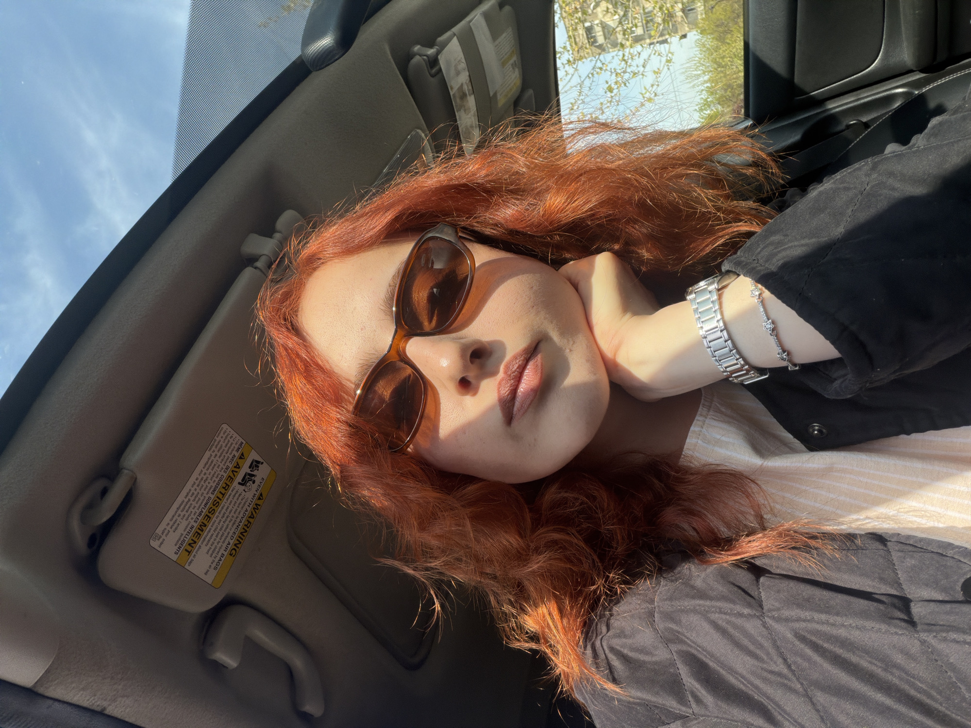 A young woman with long, flowing reddish-brown hair is captured in a selfie-style photo, taken from inside a car in Tbilisi, Georgia, during daytime. The image is oriented with the car's ceiling on the left and the car door on the right. She is wearing brown-tinted sunglasses and a black quilted jacket over a light-colored top. Her left hand, adorned with a silver watch and a silver bracelet, props up her head, with her elbow resting out of frame. Her lips are slightly parted, and her expression is neutral.
Bright sunlight streams into the car, casting noticeable shadows on her face and highlighting her hair. The car's interior features a beige sun visor with a warning label, visible in the upper part of the frame. Through the car window, a clear blue sky and green trees with visible branches suggest a sunny, outdoor environment.
The text on the sun visor warning label is clearly legible, appearing in both English and French. The English section reads: "WARNING EVEN WITH ADVANCED AIR BAGS Children can be killed or seriously injured by the air bag Never put a rear-facing child seat in the front The back seat is the safest place for children Always use seat belts and child restraints See owner's manual for more information about air bags." The French translation is also present below the English text.
