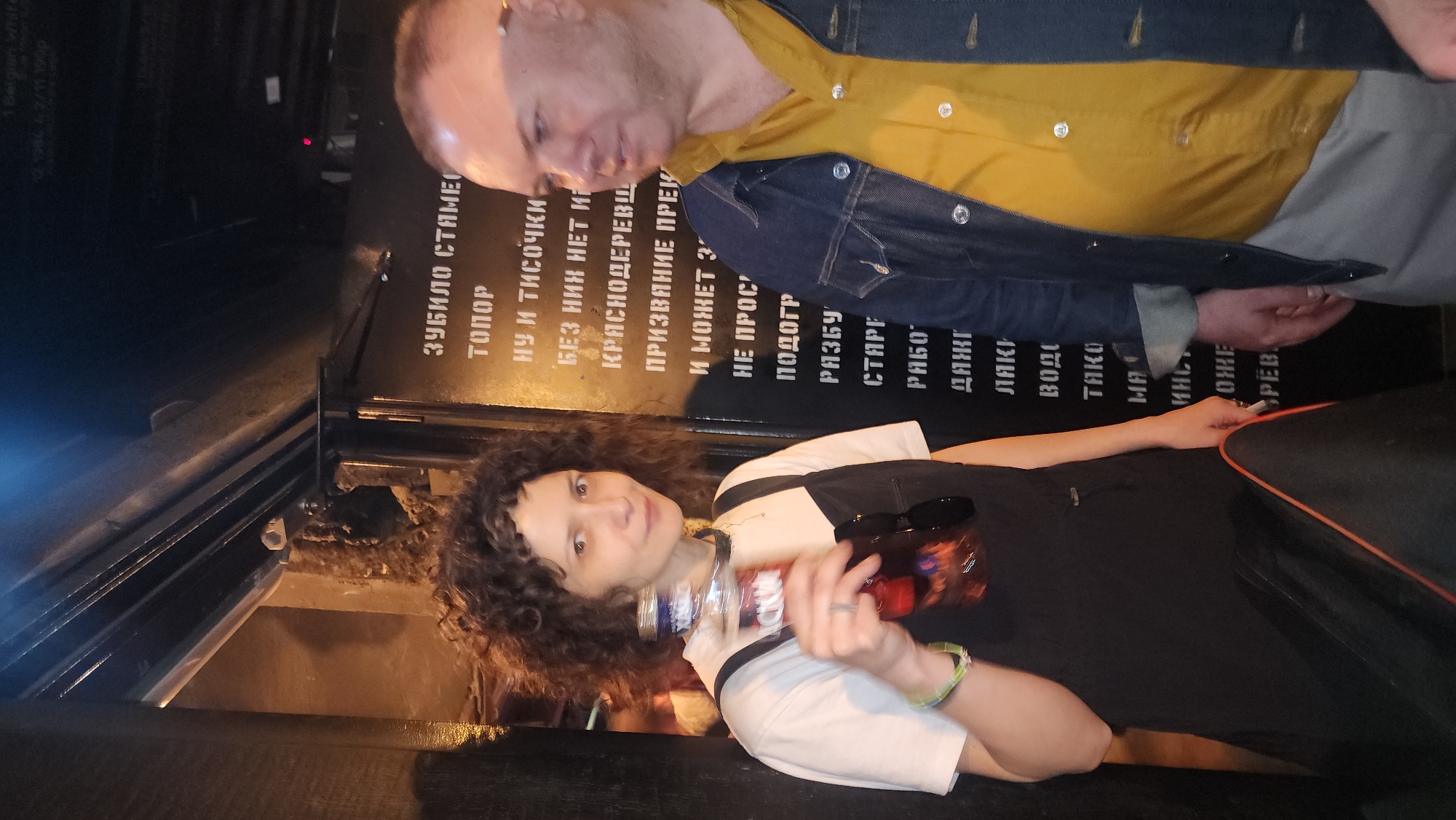 The image shows a woman with curly hair holding a bottle, possibly alcohol, with a man partially visible above her. The setting appears to be indoors, likely a bar or establishment in Moscow, Russia. This is indicated by the Cyrillic lettering on a black panel behind them, listing words related to tools and materials, likely part of the decor. The woman is wearing a black and white outfit, and the man is wearing a yellow shirt and denim jacket. The overall lighting is dim, creating an intimate atmosphere.