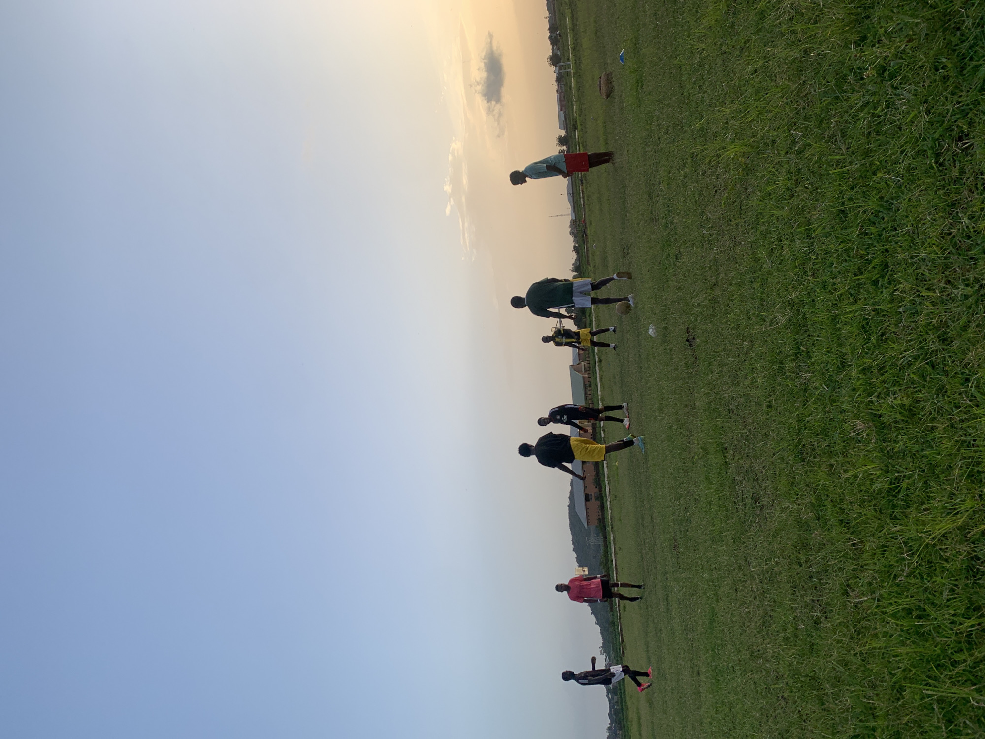 The image shows a group of young men on a grassy field, likely a football pitch, in Gwafan, Nigeria. The sky is clear with a gentle gradient from pale blue to a warm, peachy hue near the horizon, suggesting it is either early morning or late afternoon. The sunlight casts long shadows, indicating a low sun angle.
There are approximately seven individuals visible, dressed in athletic wear. Most are wearing t-shirts and shorts, with some in football jerseys and shorts. Several of them are engaged in what appears to be football training or a practice session. One person in the foreground is partially visible, wearing a black t-shirt and white shorts. Further up the field, another person in a red t-shirt and black shorts is also standing. Above them, two individuals are seen in black jerseys and yellow and black shorts respectively, with one appearing to be kicking a football. Higher still, a person in a green jersey and white shorts is visible, and another in a teal t-shirt and red shorts is performing a plank. The ground is covered in well-maintained green grass, with some sparse vegetation and distant buildings or structures visible on the horizon. The overall atmosphere appears to be one of focused activity and athletic pursuit in an open, outdoor setting.