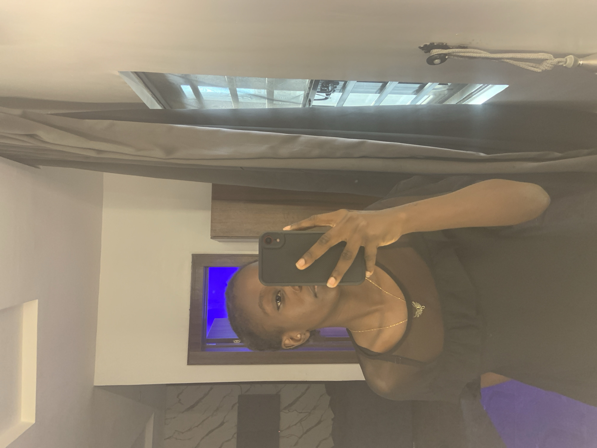 A woman in a black top is taking a selfie with a black smartphone. The selfie is taken indoors. Behind the woman is a gray curtain, a ceiling window showing a metal roof structure, and a wall with a framed window emitting blue light. The environment suggests a modern, well-lit room. The presence of these elements, although not directly indicative of Ota, Nigeria, does not exclude such a possibility. There is no specific information to pinpoint the location to Ota, Nigeria.