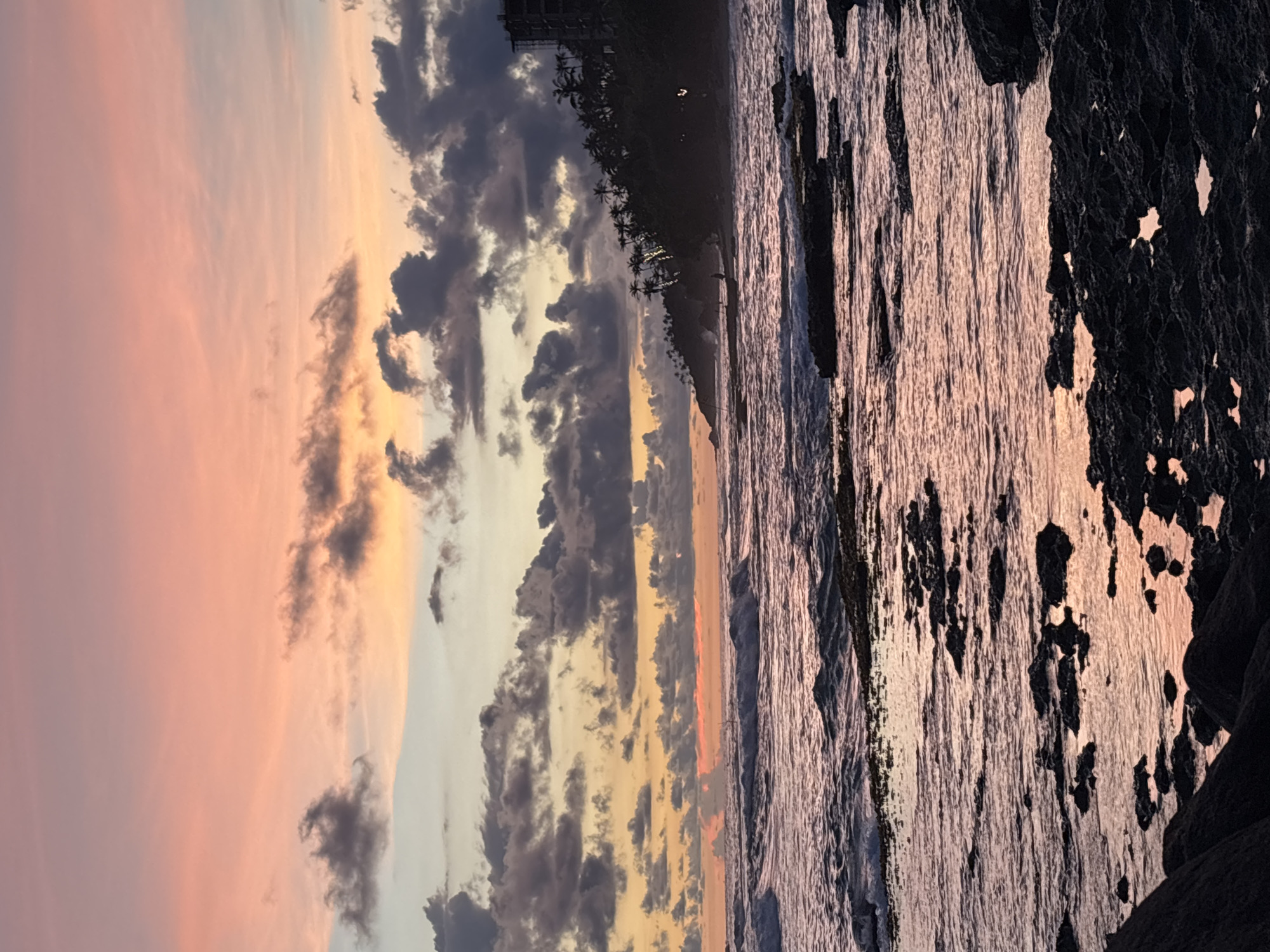 The image depicts a serene sunset scene in Thalaramba, Sri Lanka. The sky is a blend of pink, orange, yellow, and gray hues, marked by rows of clouds. The sun reflects in the ocean. Along the shoreline, palm trees are visible, lining the coast and a building in the background. Waves break against rocks at the edge of the frame.