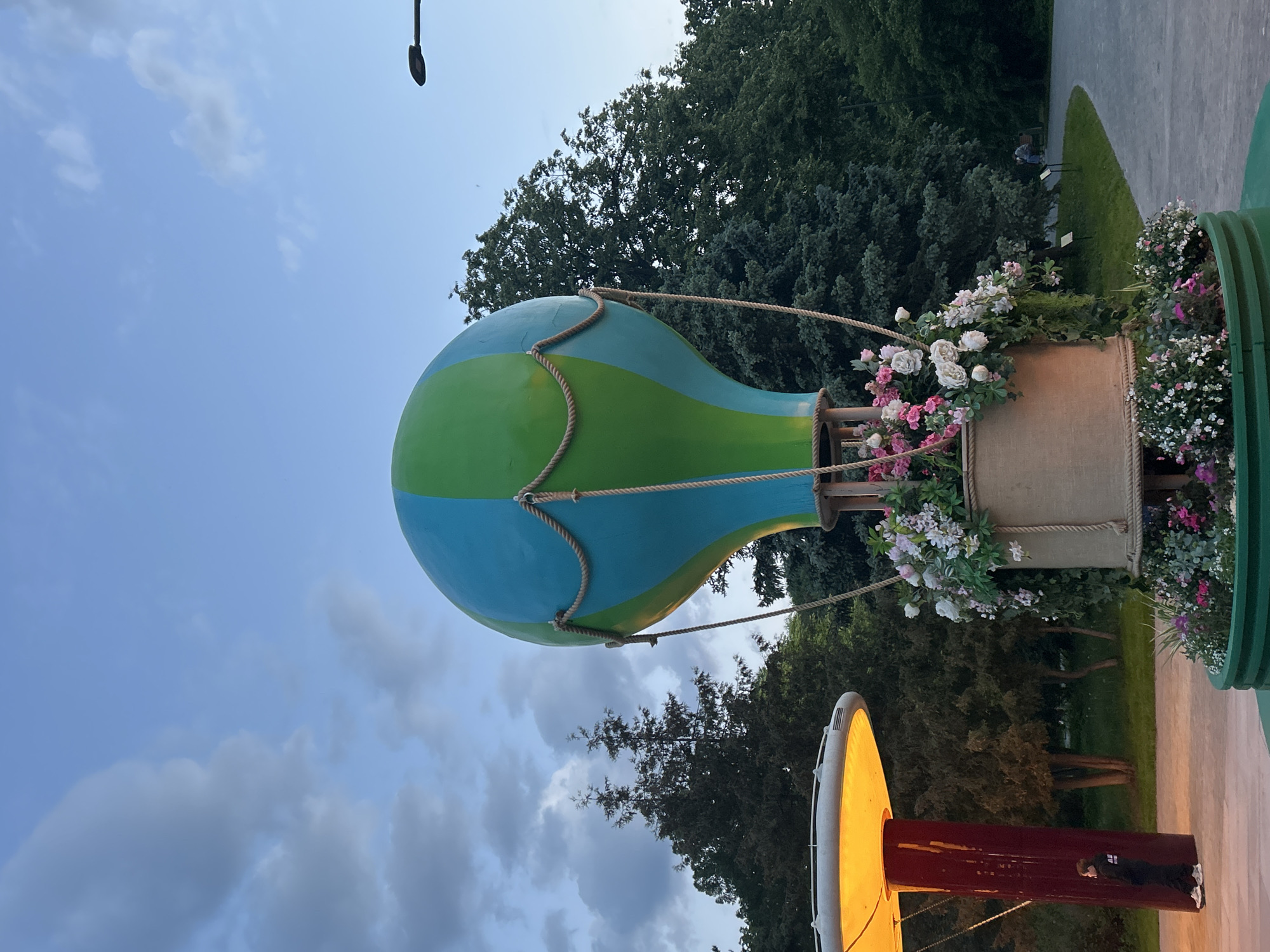 This vertical image features a large, decorative hot air balloon structure set in an outdoor park or garden scene, likely during late afternoon or early evening. The balloon itself is painted with alternating bright blue and green stripes and is adorned with thick rope detailing, giving it a realistic appearance. Below the balloon, a basket-like base, possibly made of burlap or a similar fabric, is heavily decorated with an abundance of white, pink, and purple flowers. More flowers in various hues spill from green planters around the base of the structure.
In the background, a dense expanse of dark green trees forms a natural backdrop, indicating a well-treed area. A paved path curves through a grassy section on the right, leading towards the trees. A tall, dark street light pole is visible against the sky near the top of the frame. Towards the bottom left, another prominent light fixture stands out, featuring a bright yellow, dome-shaped top that appears to be illuminated, resting on a dark red, cylindrical base. A person, dressed in dark clothing and white shoes, is standing next to this red and yellow light fixture, looking to the left.
The sky is mostly clear with some scattered, soft clouds, and the light suggests either dusk or early night, given that the artificial lights are active. The scene conveys a decorative and pleasant ambiance, likely an installation in a public space. No discernible text is visible on any of the main elements.