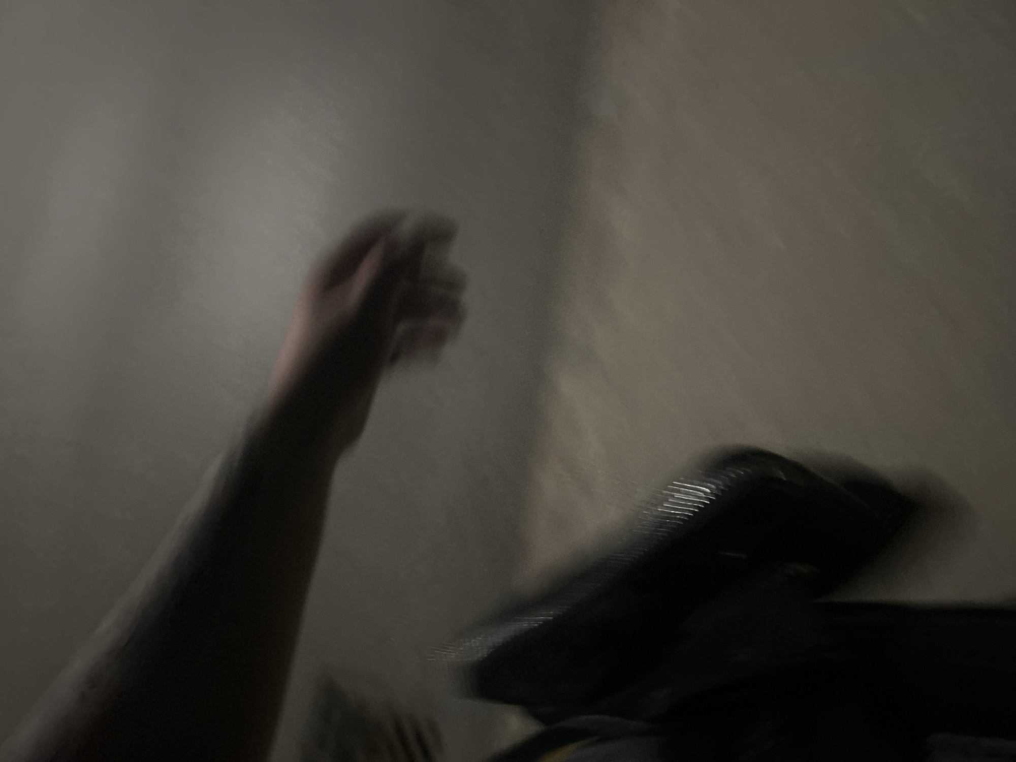 The image shows a blurry view of a person's arm and hand. The person is holding a black bag. The background is a neutral-colored wall or ground. The image appears to be taken in low light conditions, possibly at night. The blurriness suggests movement or a fast shutter speed. The location is unknown.