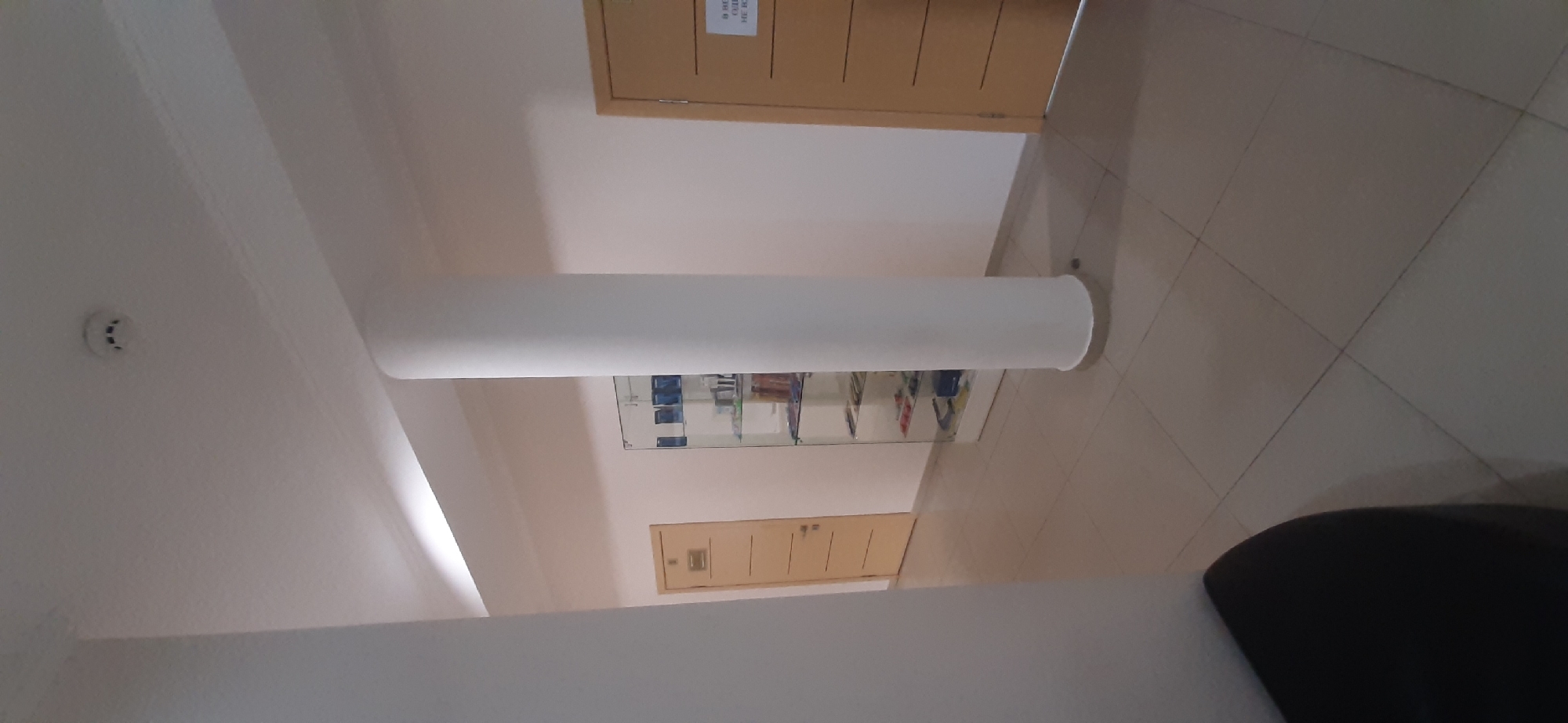 The image depicts an indoor scene, likely a hallway or common area in a building in Yessentuki, Russia. The orientation suggests the photograph was taken looking upwards and sideways, possibly from a low angle or due to the camera being held vertically.
Dominating the center of the frame is a large, cylindrical white pillar, which appears to be a structural element. Below the pillar, a glass display cabinet is visible, filled with various items that are difficult to discern clearly but seem to include bottles or containers. To the right of the pillar, a closed wooden door with horizontal paneling is present. Above this door, a smaller sign is affixed, with some text visible, though it is mostly illegible.
Further to the left, near the top of the image, another wooden door is visible. Above the pillar and to the left, a circular smoke detector is mounted on the wall. The floor is tiled with large, square, light-colored tiles, laid in a grid pattern.
The lighting appears to be artificial and even, suggesting it is daytime or the interior is well-lit. There are no people visible in the image. The overall impression is one of a clean, modern, and functional interior space. The camera's perspective creates a somewhat abstract composition, emphasizing the architectural elements and the display cabinet.