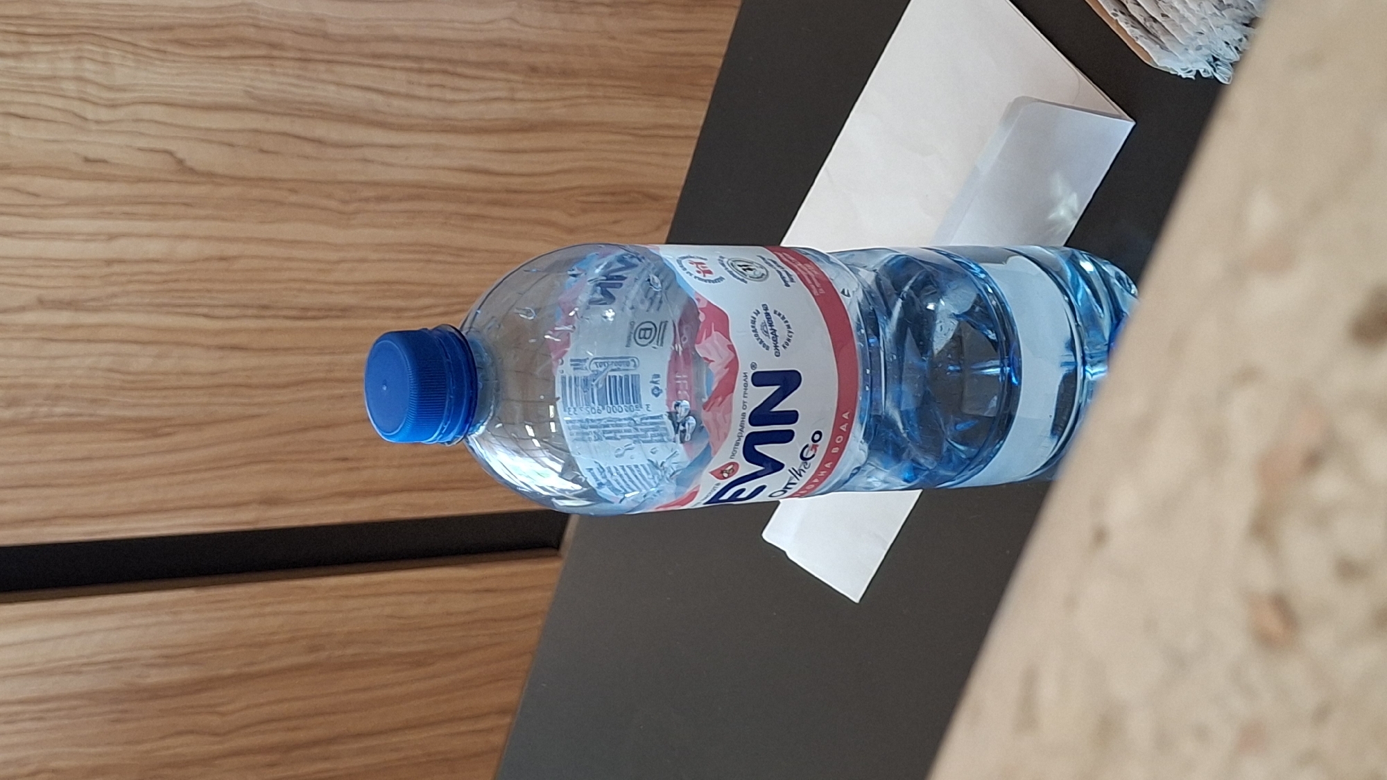 A clear plastic bottle of "EVIN On The Go" drinking water, with a blue cap, stands upright on a dark surface. The bottle is partially filled with blue-tinted water. A white folded piece of paper is positioned behind the bottle. The background consists of a wooden wall with vertical paneling. The scene appears to be indoors, possibly in a commercial establishment or a hotel in Nesebar, Bulgaria.