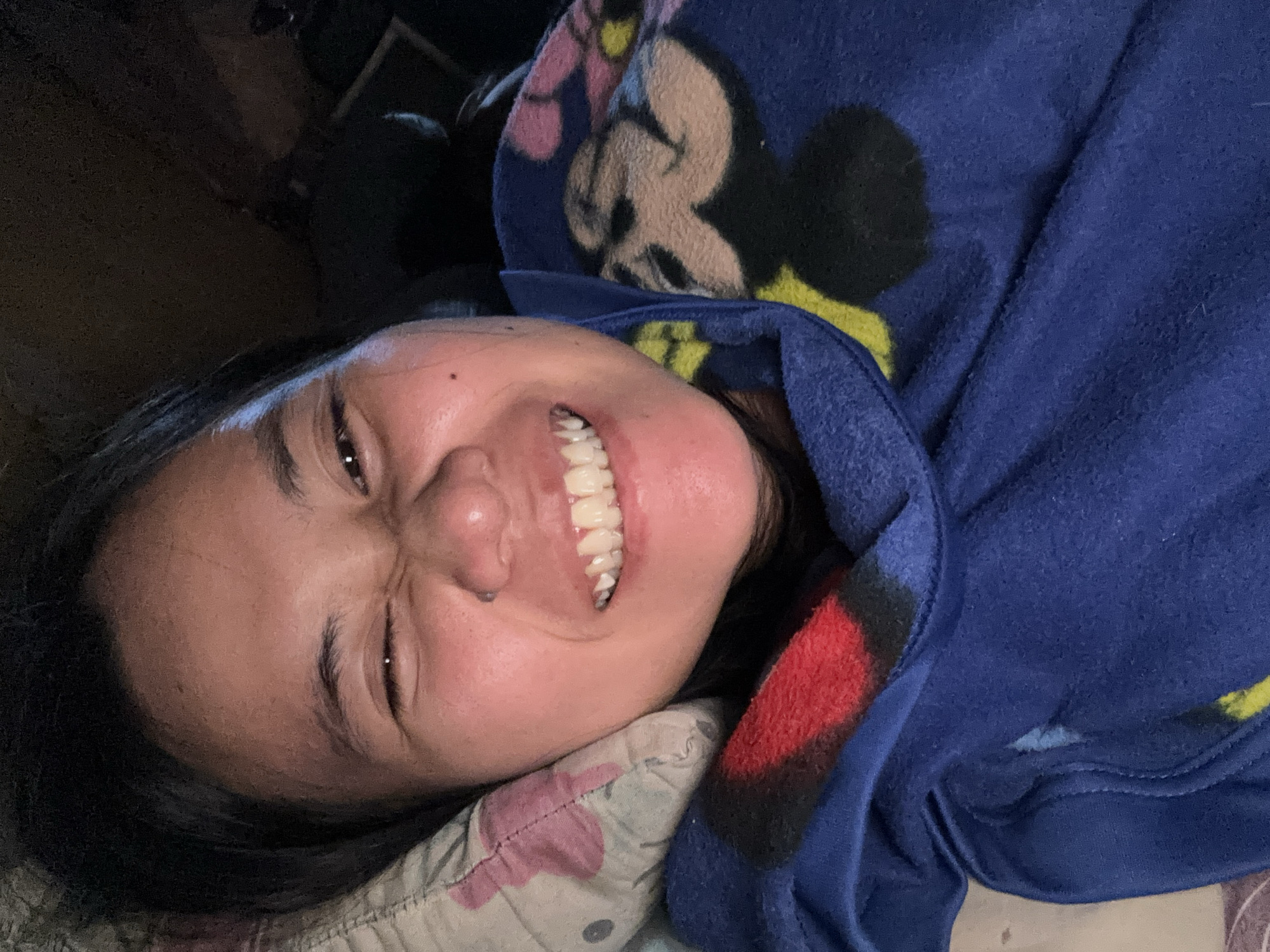A young woman is lying in bed, wrapped in a blue blanket with a Mickey Mouse pattern. She is smiling and winking at the camera. The background is dark and out of focus. The image was taken in East Jakarta, Indonesia.