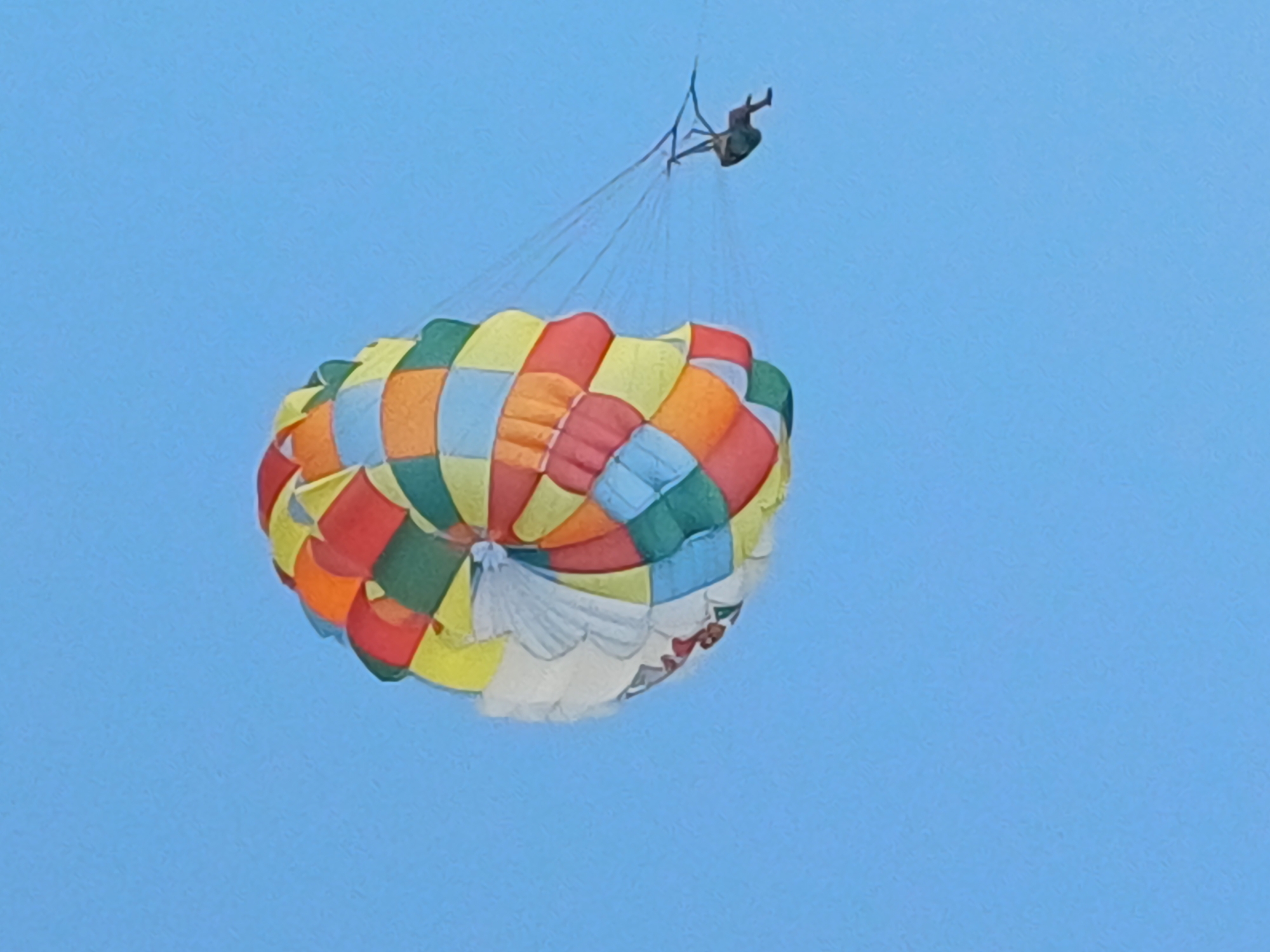 The image captures a parasailer against a clear blue sky, likely over Sochi, Russia. The parasail is a vibrant, multi-colored canopy with sections of red, orange, yellow, green, and blue squares arranged in a circular pattern. The parasail is inflated and appears to be in mid-flight, with thin ropes connecting it to a person who is suspended above. The person is a dark silhouette against the bright sky, wearing what appears to be a harness and in a seated or kneeling position. The scene suggests an activity or event taking place outdoors, likely on a sunny day given the bright sky and the illuminated parachute. The overall impression is one of leisure and adventure, characteristic of activities enjoyed in a coastal or resort area like Sochi. There are no discernible location cues beyond the provided context, and no visible text. The image is a close-up, focusing on the parasail and the person, with the vast expanse of the sky as the background.