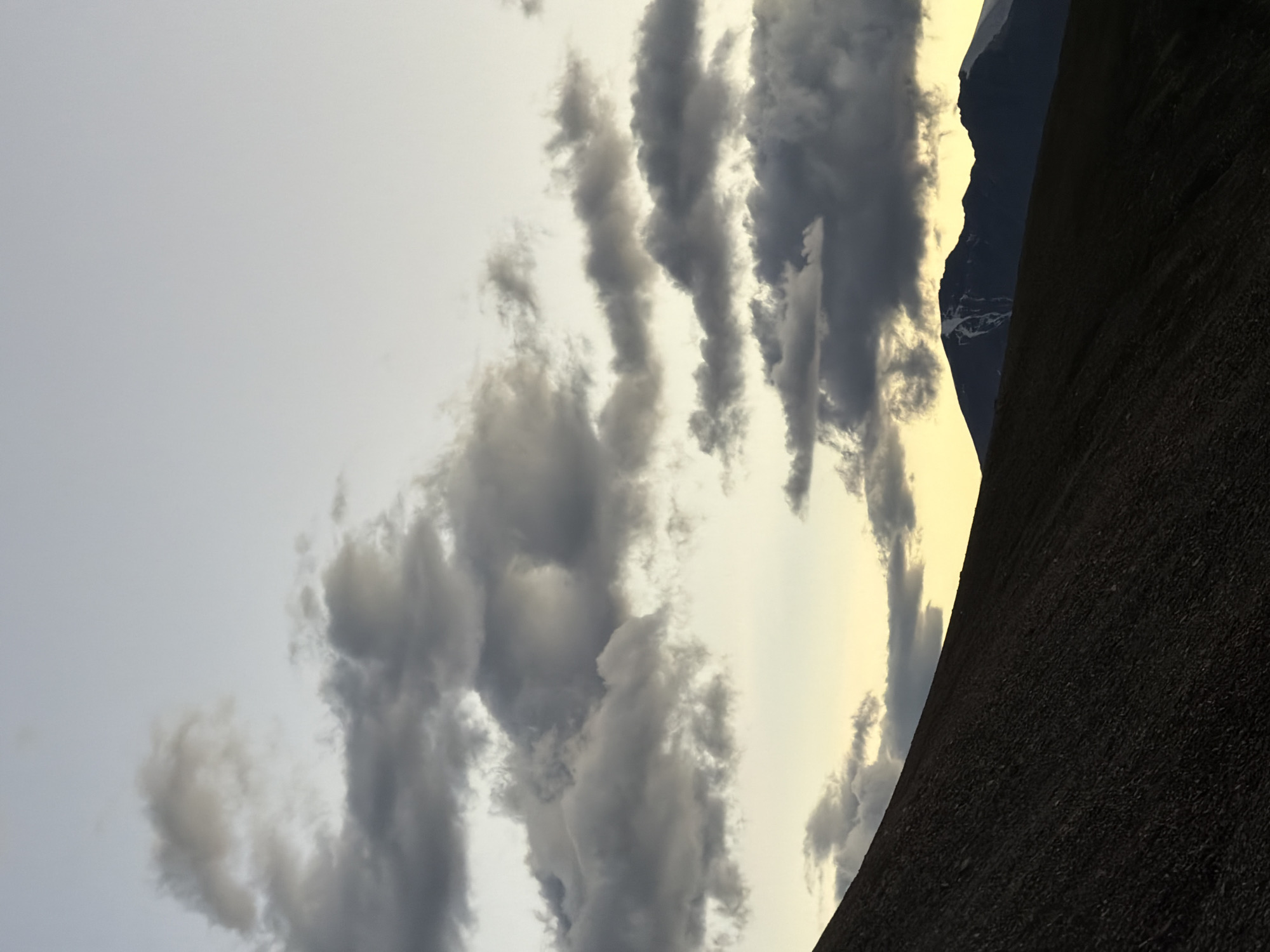The image depicts a twilight scene in a mountainous landscape. In the foreground, a dark, sloped terrain dominates the left side of the frame, its texture suggesting a rocky or gravelly surface. The middle ground features the silhouette of distant mountains against a pale yellow sky, with patches of snow visible on their peaks. Dark, scattered clouds drift across the upper portion of the image, set against a lighter, pale blue sky. The overall impression is one of natural, undeveloped wilderness. The specific location in the city of Unknown, India, is not discernible from the image itself.