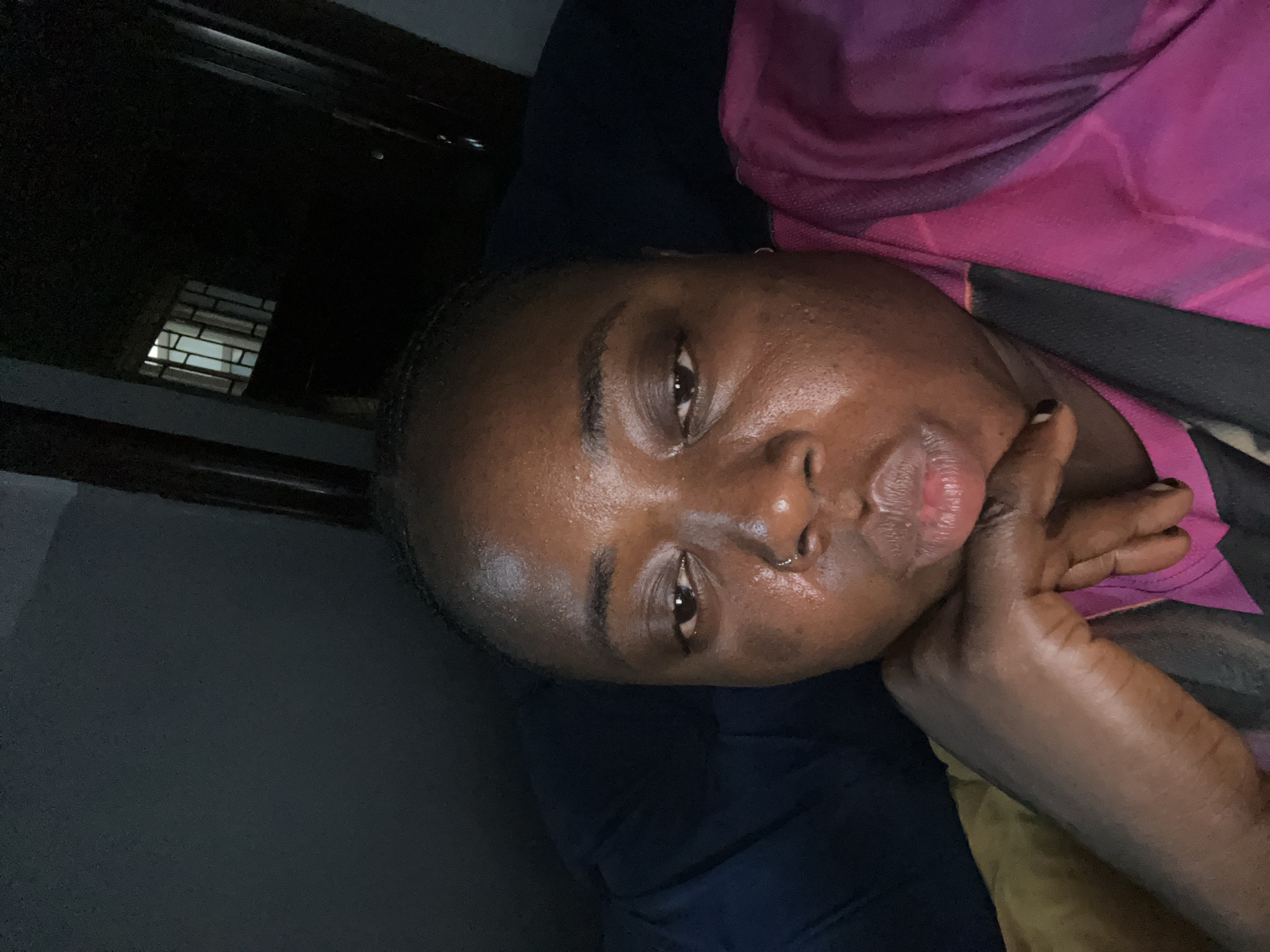 A person is lying down with their head resting on their hand, in what appears to be an indoor setting. The individual has dark skin, short dark hair, and a piercing in their nostril. They are wearing a pink top and a black strap is visible near their chest. The background consists of a dark wall and possibly a window with bars. Given the constraint to locate the image in Kubwa, Nigeria, it's not possible to confirm that the image originated there, but the overall setting is consistent with residential areas in Kubwa. There is no additional evidence available to indicate this is from Kubwa.