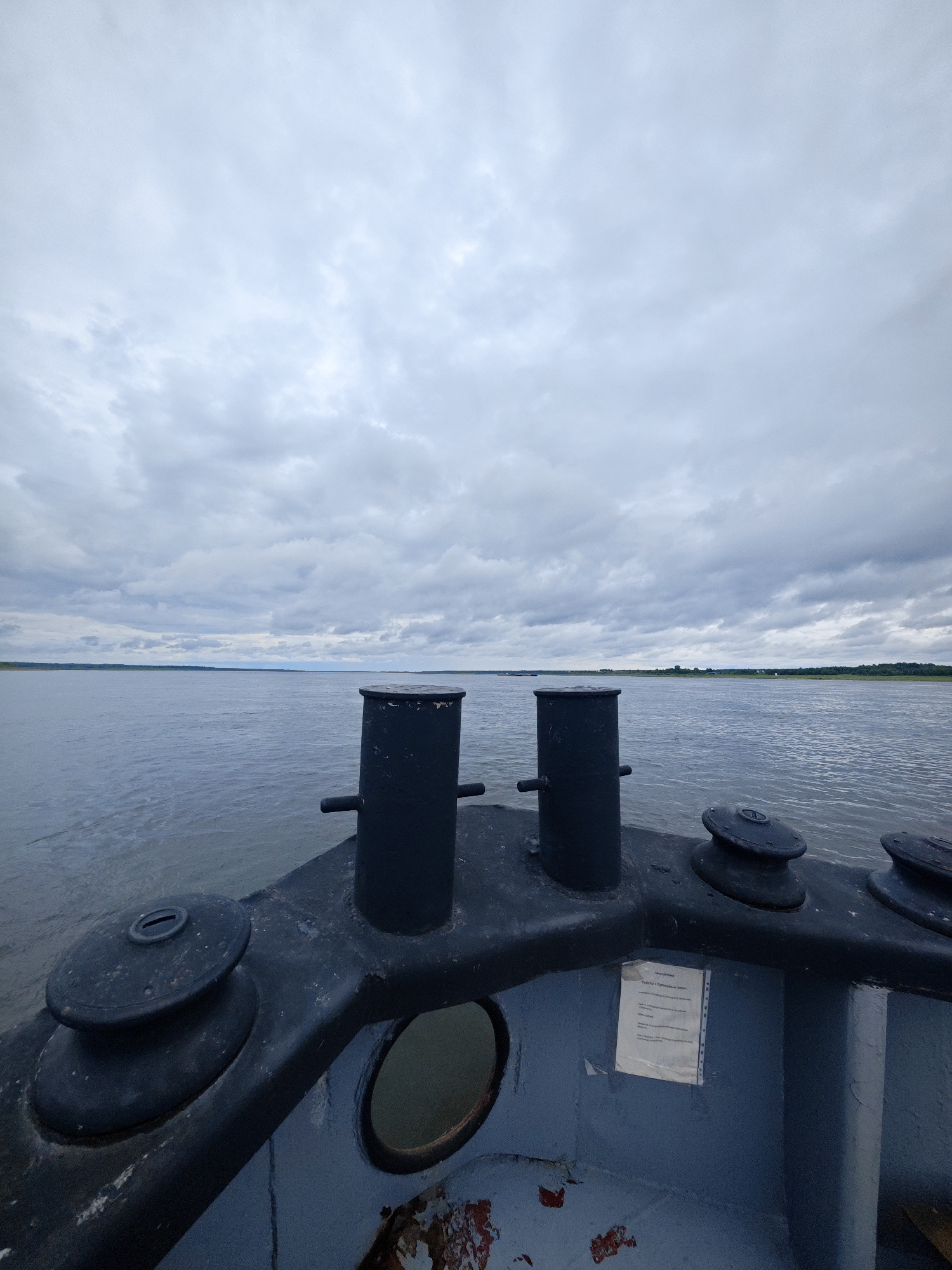 The image depicts a forward view from a boat on a large, calm body of water, likely a river, under an overcast sky. The foreground of the image is dominated by the deck of the boat, featuring two dark, cylindrical bollards and two boat winches. The deck appears weathered, with peeling paint revealing rust and primer in places. A circular porthole is visible on the side of the boat's structure. In the middle ground, the expanse of the water stretches to the horizon, where a low, tree-lined shore is visible under the cloudy sky. The overall impression is one of a quiet, possibly industrial, maritime setting. Given the context of Zotino, Russia, this scene likely represents a view from a vessel navigating the Yenisei River, a major waterway in Siberia.