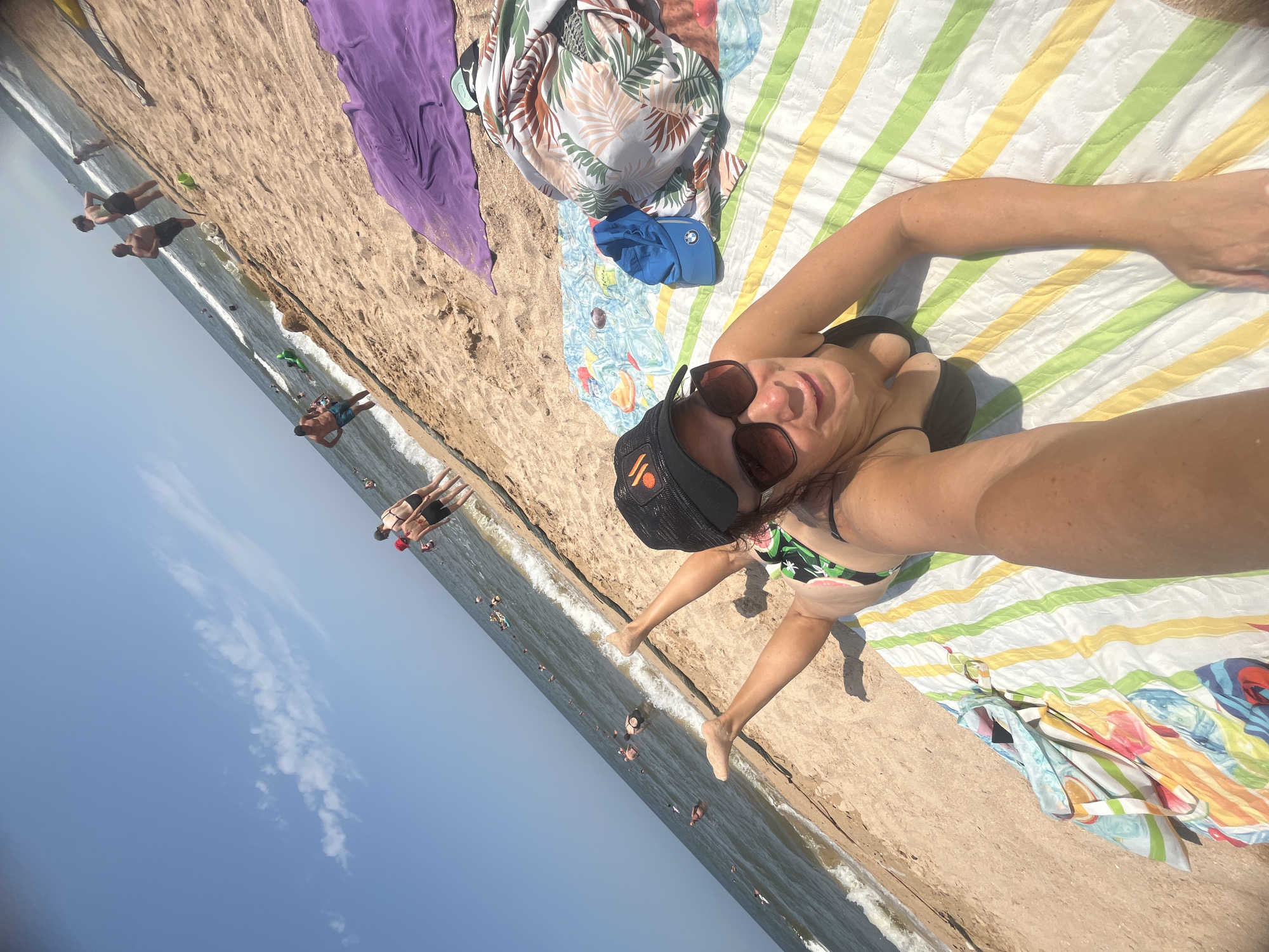 A woman is lying on a striped beach towel on a sandy beach in Golubitskaya, Russia. She is wearing sunglasses and a black baseball cap. The sea is in the background with several people swimming and standing in the water. There are also beach towels and other beach accessories scattered on the sand. The sky is clear and blue with a few scattered clouds. The image is taken from a high angle, looking down at the woman.