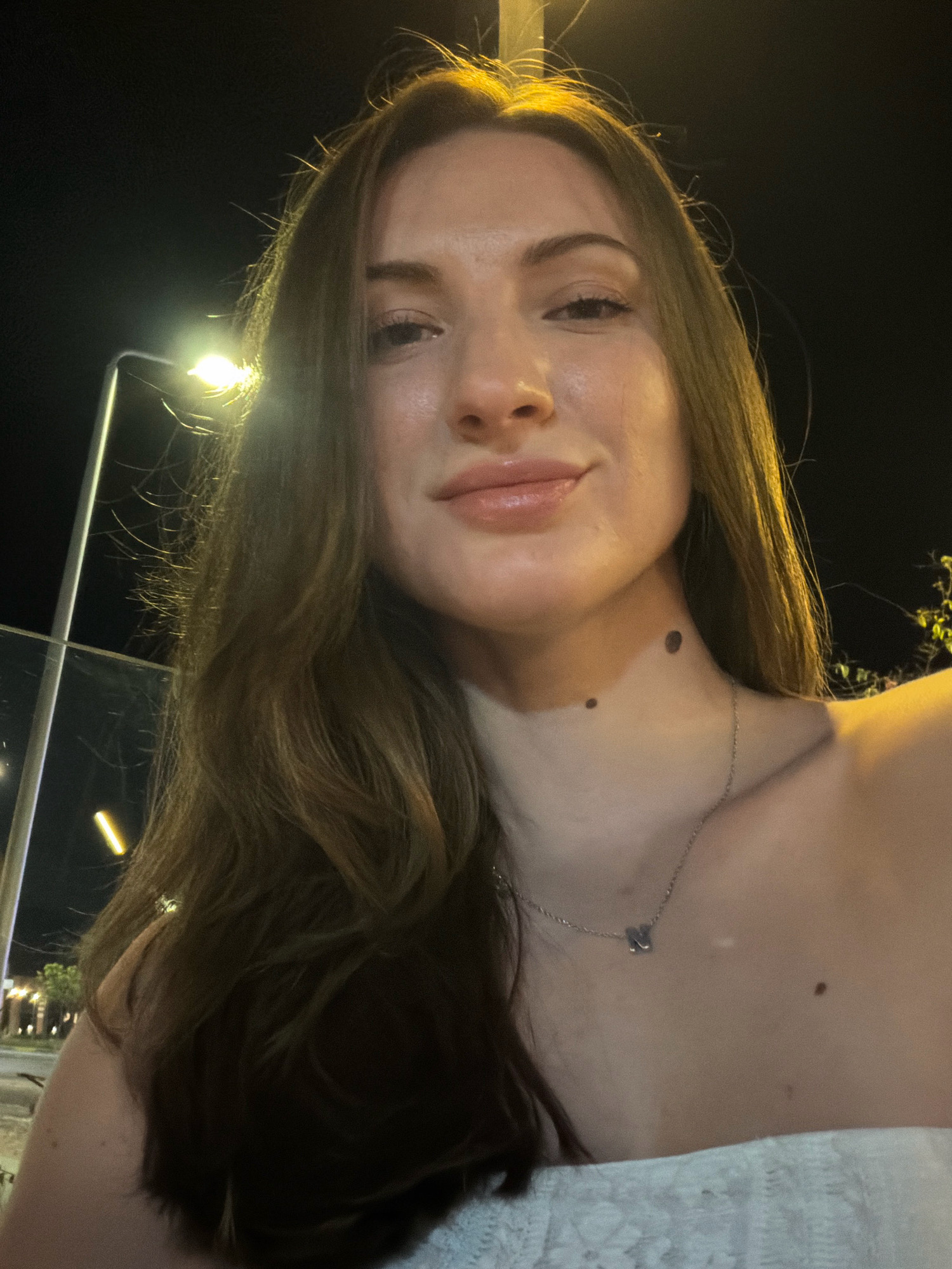 A young woman with long brown hair and fair skin is pictured in a close-up, self-taken photograph at night. She is looking directly at the camera with a slight smile. She is wearing a white strapless top and a silver necklace with a small butterfly pendant. There are two moles on her neck and one on her chest. The background is dark, with a street light visible on the left side of the frame, suggesting an outdoor setting in a city. The overall lighting is dim, with the street light providing a warm glow on the woman's face and hair. The location is identified as Sharm El Sheikh, Egypt.