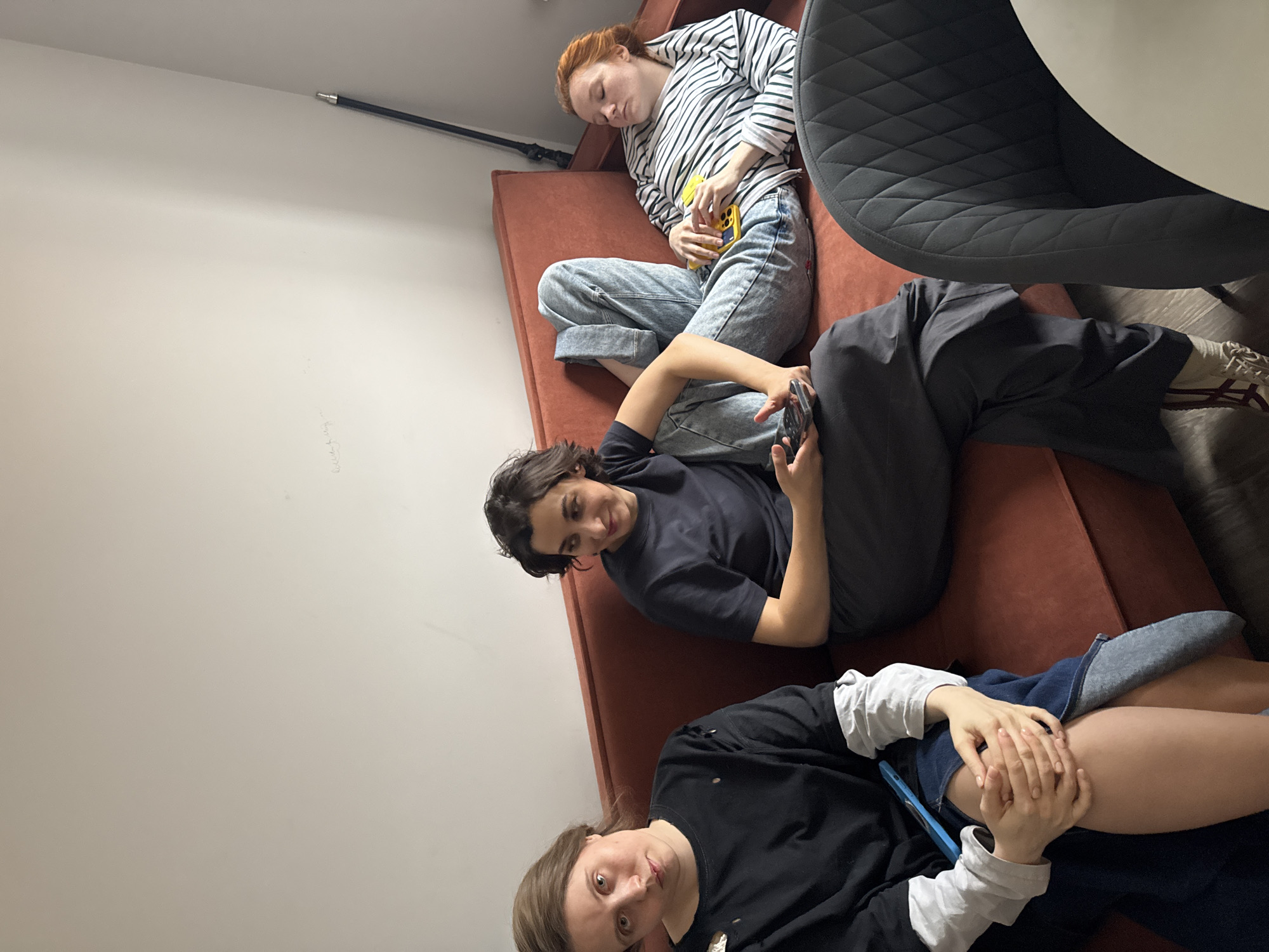 The image depicts three individuals on a rust-colored sofa against a light gray wall. An auburn-haired person is lying at the top of the image, wearing a striped shirt and light-wash jeans, with a yellow cell phone in hand. Adjacent to this individual is a person in a dark t-shirt, with dark hair, holding a cell phone and looking towards the camera. At the bottom of the image, a person with light brown hair is wearing a black long-sleeved shirt with white sleeves underneath, holding a blue cell phone. A black rod is visible near the top of the wall. Part of a black chair and a white table are in the top right of the image. The location is Unknown, Unknown.