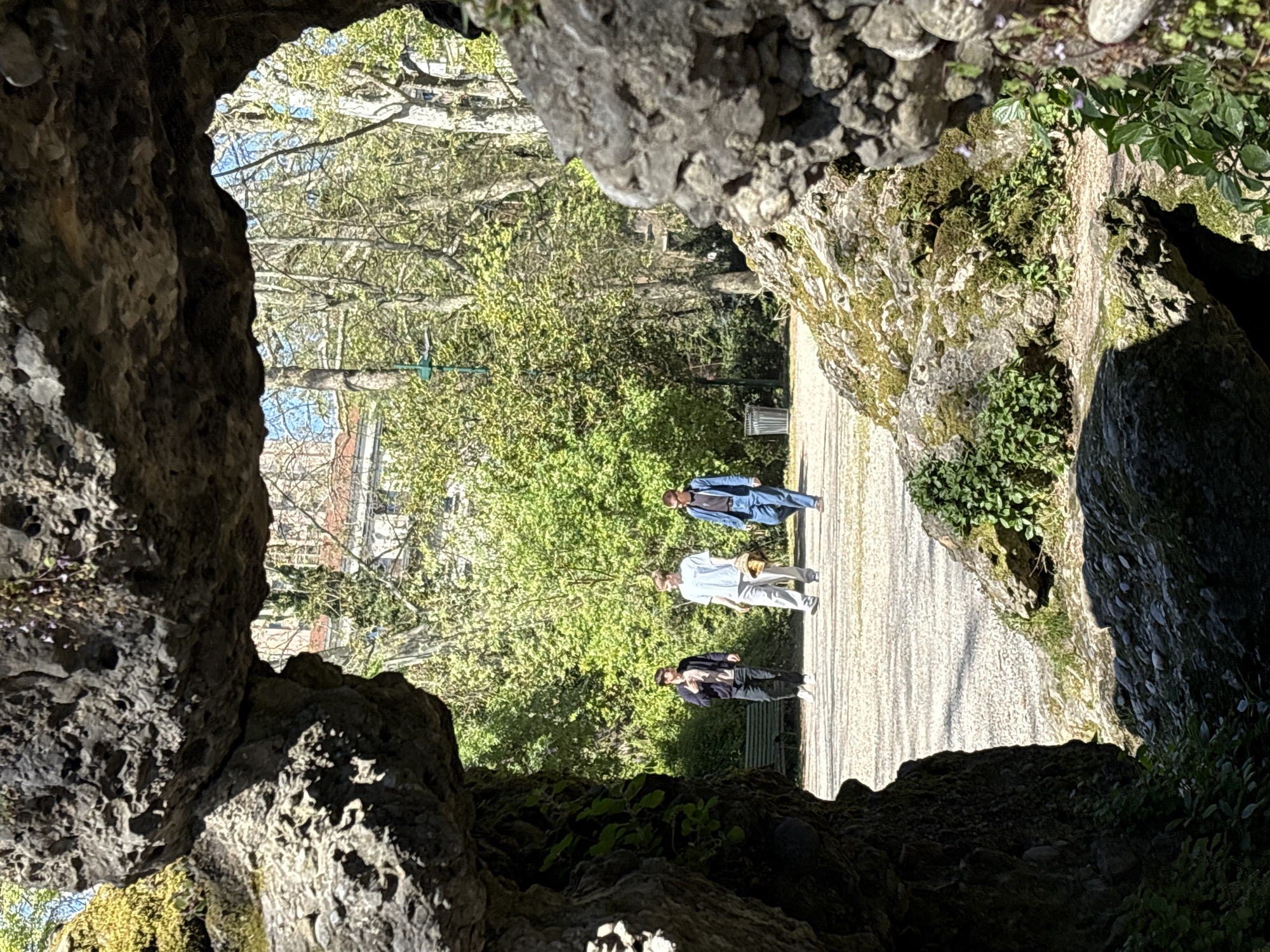 The image depicts a park scene viewed through a rocky opening, possibly a cave or grotto. Three people are walking along a gravel path away from the viewer. The person closest to the camera is wearing dark pants and a dark jacket. The middle person is wearing light-colored pants and a light-colored shirt, and carrying a jacket over their shoulder. The person furthest away is wearing blue jeans and a blue jacket. A green park bench is seen near the person closest to the camera. Trees and greenery line both sides of the path, creating a tunnel effect. Some buildings are visible in the distance. This is located in Sempione Park in the city of Milan, Italy.