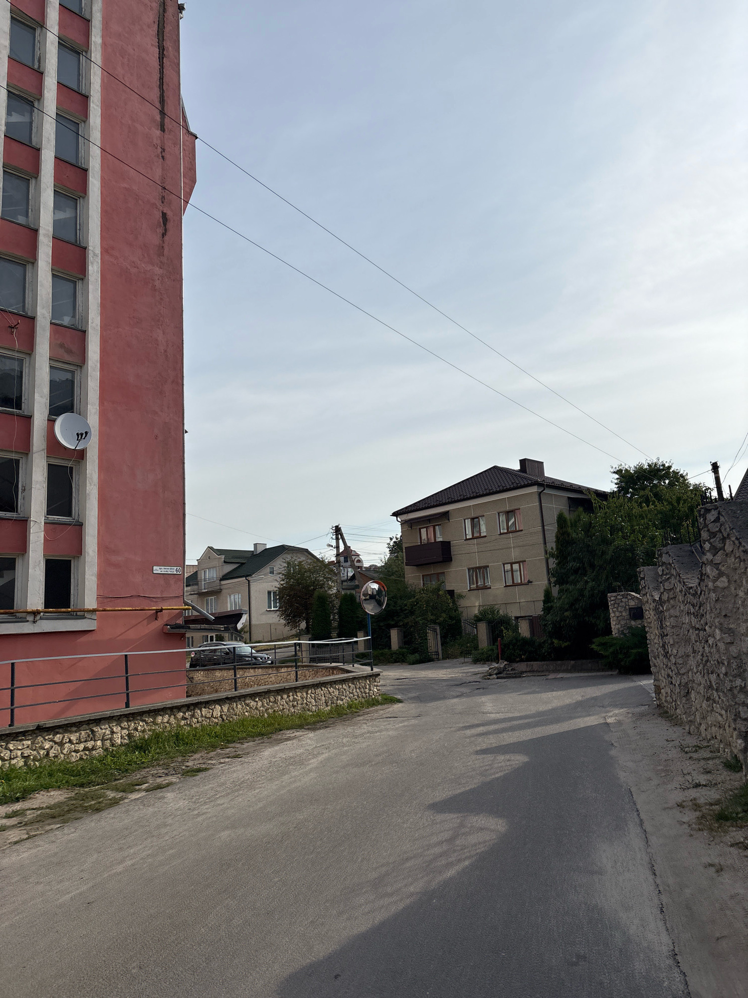The image shows a street scene in Ternopil, Ukraine, with a large, faded pink building on the left. This building has a facade of white vertical and horizontal lines, with rows of windows. A satellite dish is mounted on the side of the building. A metal railing runs along a low, stone-faced wall in front of the building, with some grass growing between the stones.
The street itself is paved and slopes gently upward towards the right. On the right side of the street, there is a wall made of rough-hewn stones. Beyond the stone wall, there are trees and a building with a dark, angled roof, several windows, and balconies. Further in the distance, a traffic mirror is visible, reflecting a portion of the street and buildings. The sky is overcast with a pale blue hue. The overall impression is of a residential or mixed-use urban area.