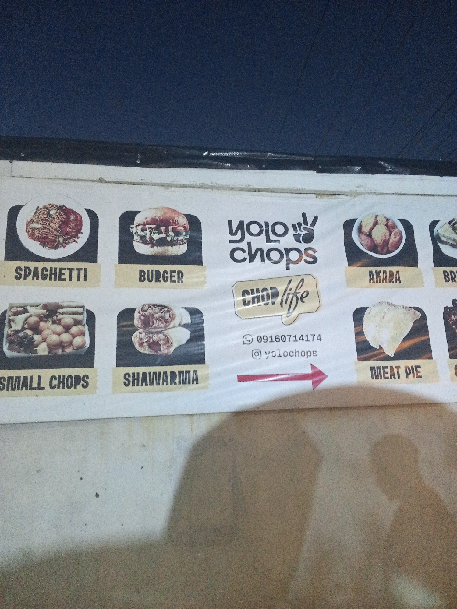 Here is a forensic description of the image:
The image shows a sign for "YOLO Chops," a food vendor. The sign features images of various menu items including spaghetti, burger, akara (bean cakes), and meat pie, as well as "small chops" and shawarma. Contact information consisting of a phone number (09160714174) and an Instagram handle (@yolochops) are on the sign as well. The background is a dark blue sky, suggesting the image was taken at night or in low light. The sign appears to be attached to a wall, which is in a state of disrepair, with a rough, textured surface. There are indistinct shadows of people in the foreground. The location, Ado, Nigeria, is not explicitly stated in the image, however, it is likely, based on the menu items, that the location is in West Africa.