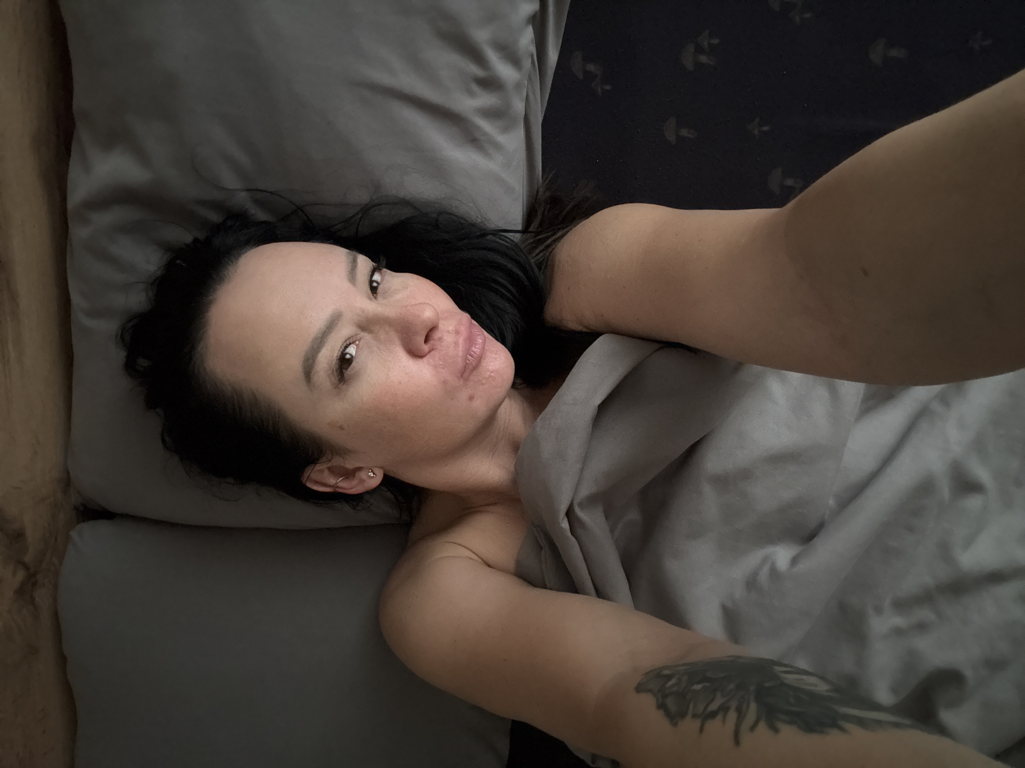 A person is lying in bed, angled toward the viewer and looking at the camera. The person has dark hair and light skin, with a tattoo visible on their upper arm. They are partly covered by a grey blanket, and a grey pillow is under their head. The bedclothes and pillowcase appear to be a similar shade of grey. The background reveals a dark-colored headboard with a repeating pattern. The image does not offer sufficient details to infer the location as Chaikovsky, Russia.